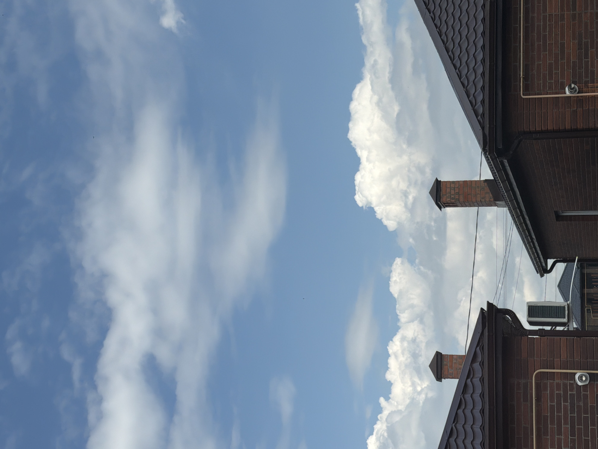 The image shows a view of the sky with clouds, above the rooftops of brick houses in the city of Krasnyi Krym, Russia. Two brick chimneys are visible extending from the dark brown tiled roofs. An outdoor air conditioning unit is mounted on the side of one of the brick houses. The sky is blue with scattered white cumulus clouds. The scene appears to be during daylight hours.
