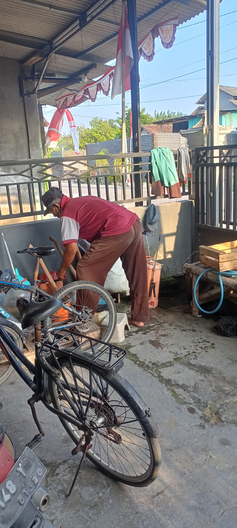 In Gempolklutuk, Indonesia, an elderly man wearing a maroon shirt with a white collar and brown pants, and a black cap is bending over, engaged in an action with a bicycle. He is standing on a concrete floor in what appears to be an outdoor shelter with a corrugated metal roof supported by metal beams. Various items are visible in the cluttered environment, including multiple bicycles, one of which is in the foreground with a black frame and a rear rack, another partially visible to its left, and a third with its rear wheel and chain mechanism prominently displayed. To the right of the man, there is an orange bucket, a small white stool, and a wooden crate. Laundry items, including a green garment, are hanging on a railing behind him. Indonesian flags and decorative banners are strung across the shelter, suggesting a festive occasion. The scene is illuminated by natural sunlight, casting shadows on the ground.