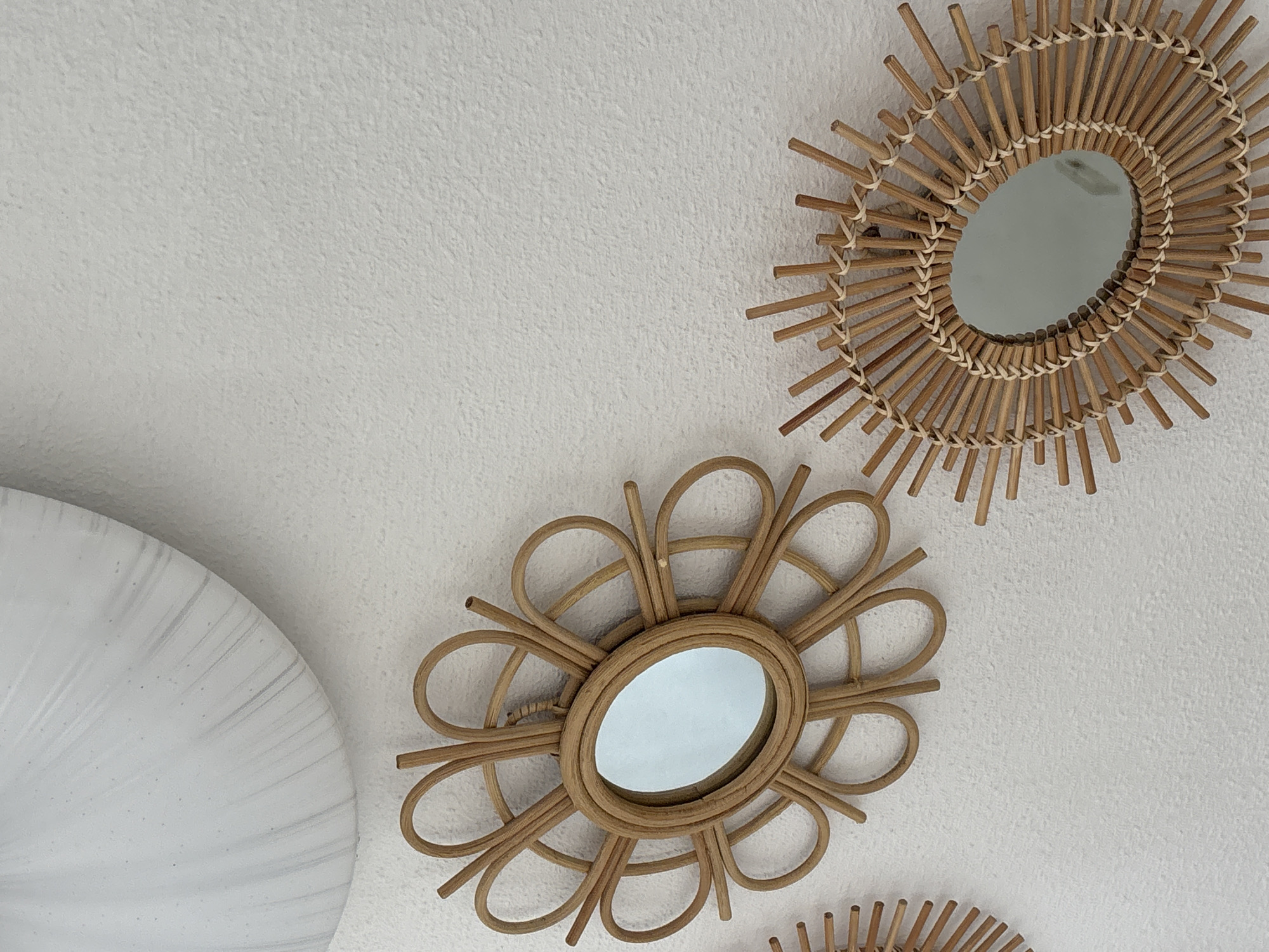 The image shows a white wall with three decorative mirrors. One large, white disc-shaped object is visible in the top left corner of the image. The other mirrors are round or oval and have a woven rattan frame. Two of the rattan mirrors are fully visible and are in focus, one with a flower-like design and the other with a sunburst design. Another rattan mirror is partially visible on the left side of the image. The wall has a textured finish. There are no people or animals in the image. The scene is an interior, likely a home or decorative space. The location is not specified in the image or accompanying information.