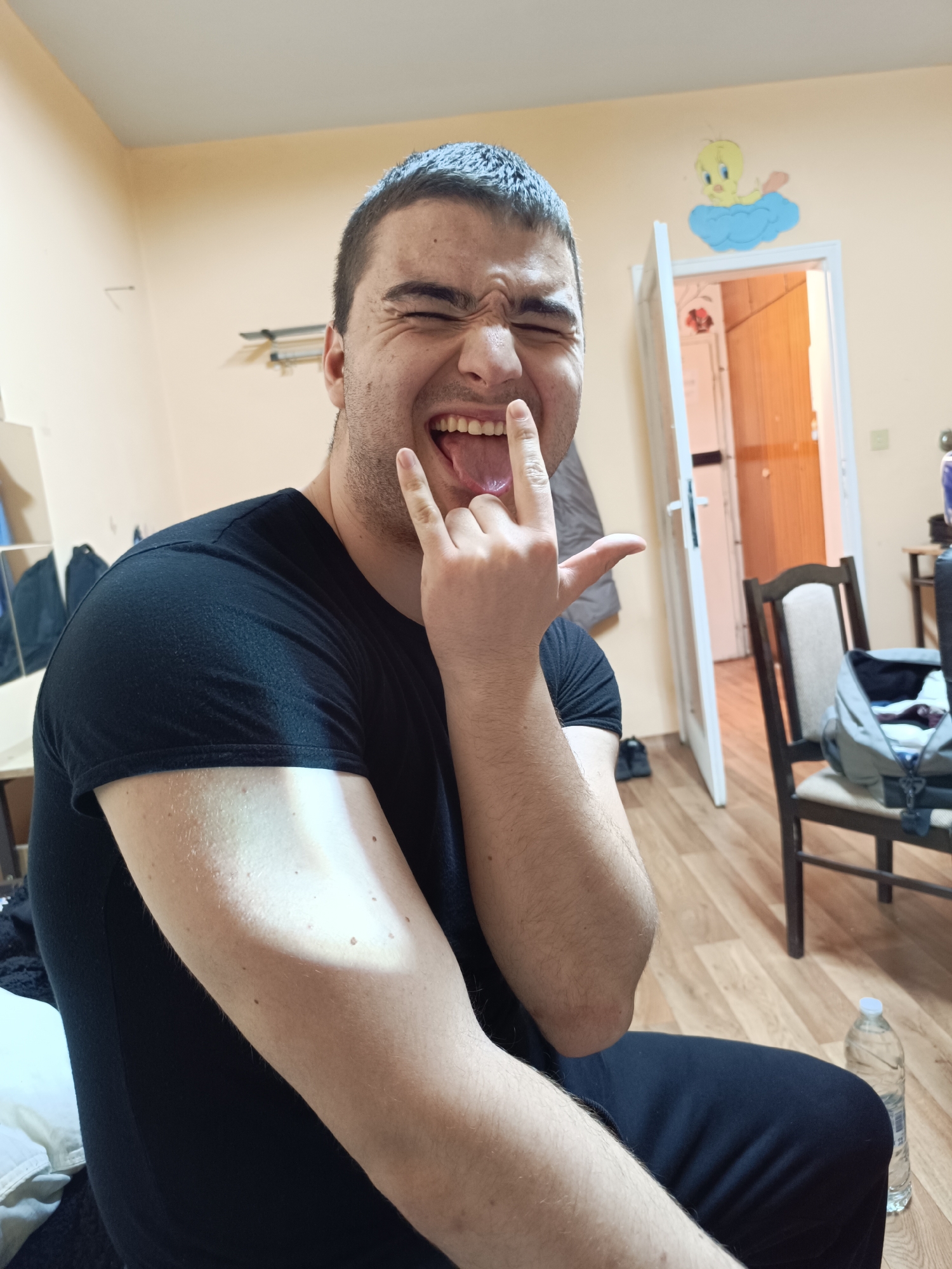 A young man is seated indoors, making a "rock on" hand gesture. He is wearing a black t-shirt and black pants. His expression is one of playful distortion, with his eyes closed and tongue sticking out. The background includes a door, a chair with a bag on it, and a water bottle on the floor. A Tweety Bird decal is visible above the door. The location details are insufficient to accurately identify a location within Sofia, Bulgaria.