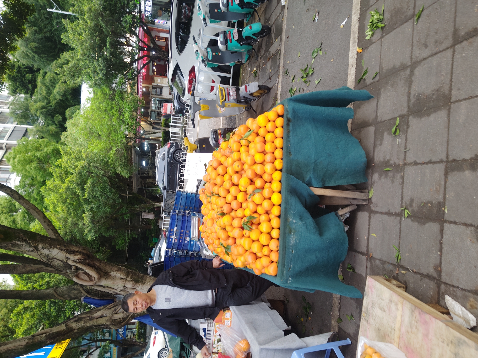 A vendor stands behind a makeshift fruit stand on a sidewalk in the Chongqing High-tech Zone, China. The stand is covered with a blue cloth and piled high with oranges. The vendor, an Asian man with short dark hair, is wearing a gray t-shirt and a black jacket. Blue plastic crates are stacked behind the table. There are motorized scooters and cars parked behind the table. A large tree is to the left of the man. Buildings are visible in the background. The sidewalk is made of gray stone blocks.