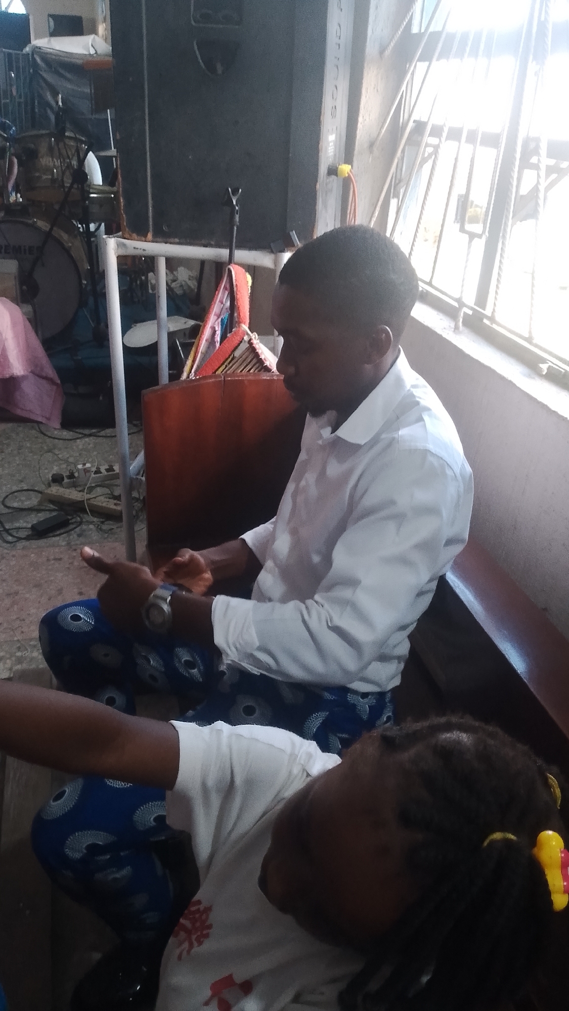 The image captures an indoor scene, likely within Warri, Nigeria, focusing on two individuals. In the foreground, a person is seen from the shoulders down, wearing a white t-shirt with a red graphic design and blue patterned trousers. Their hair is styled in braids and adorned with yellow and red hair accessories. To the left and slightly behind, an adult male is seated, wearing a white collared shirt and blue patterned trousers. He is looking down and appears to be interacting with his hands, one of which is adorned with a wristwatch. The background reveals a large black speaker and parts of a drum set, suggesting a music or performance-related setting. The overall environment appears to be dimly lit, with light filtering in from a window or opening on the right.