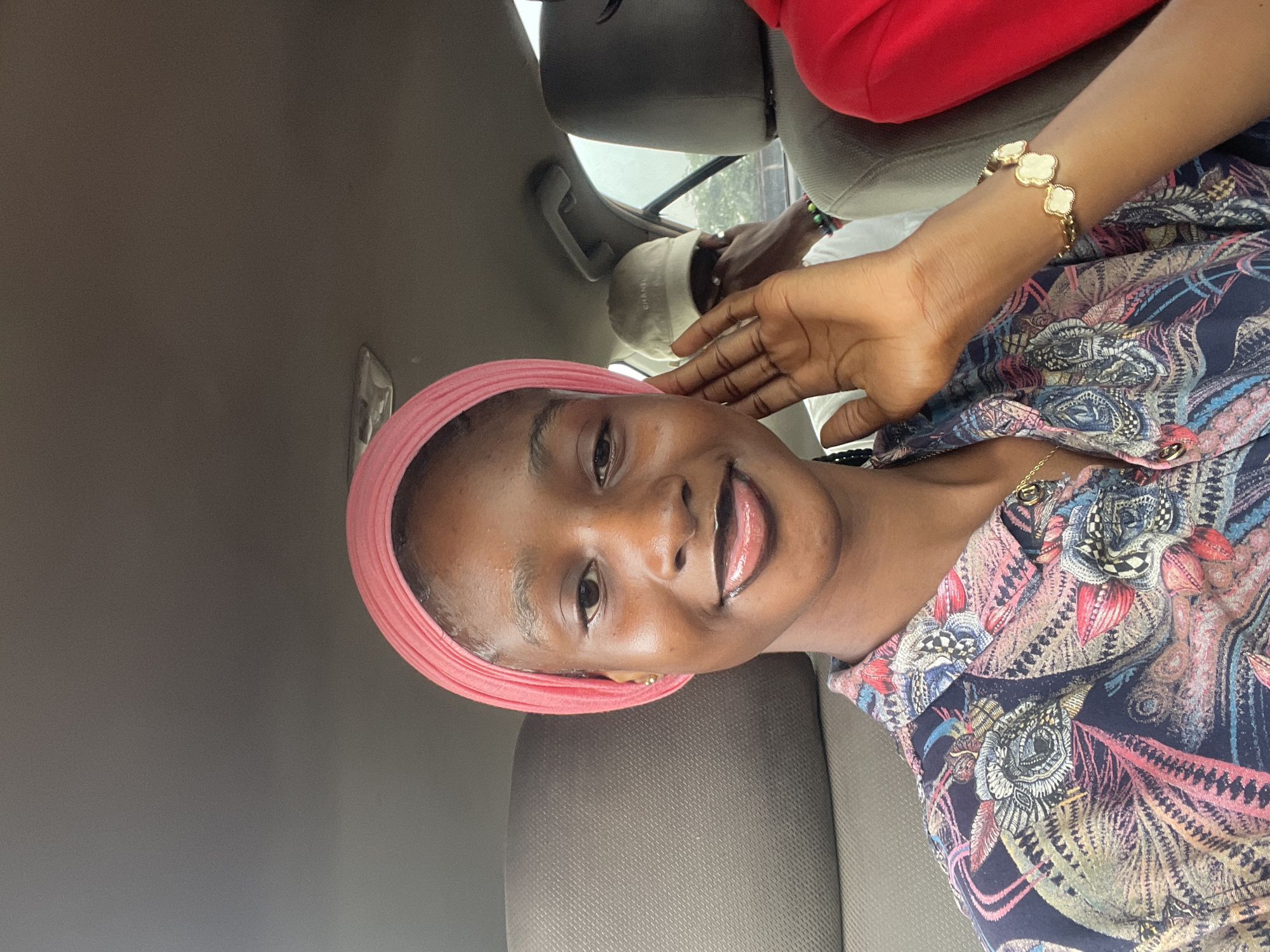 The image shows a young woman with a pink headscarf, wearing a colorful patterned shirt, taking a selfie in a car. Her right hand is raised near her face. In the background, a man wearing a beige baseball cap is visible in the passenger seat. Another person in a red top is partially visible in the rear passenger seat. The interior of the car appears to be a standard sedan. The image is likely taken during the daytime, as suggested by the lighting conditions inside the vehicle. The presence of what appears to be urban scenery outside the car windows, including trees and buildings, indicates the car is in a populated area, possibly in Abuja, Nigeria.