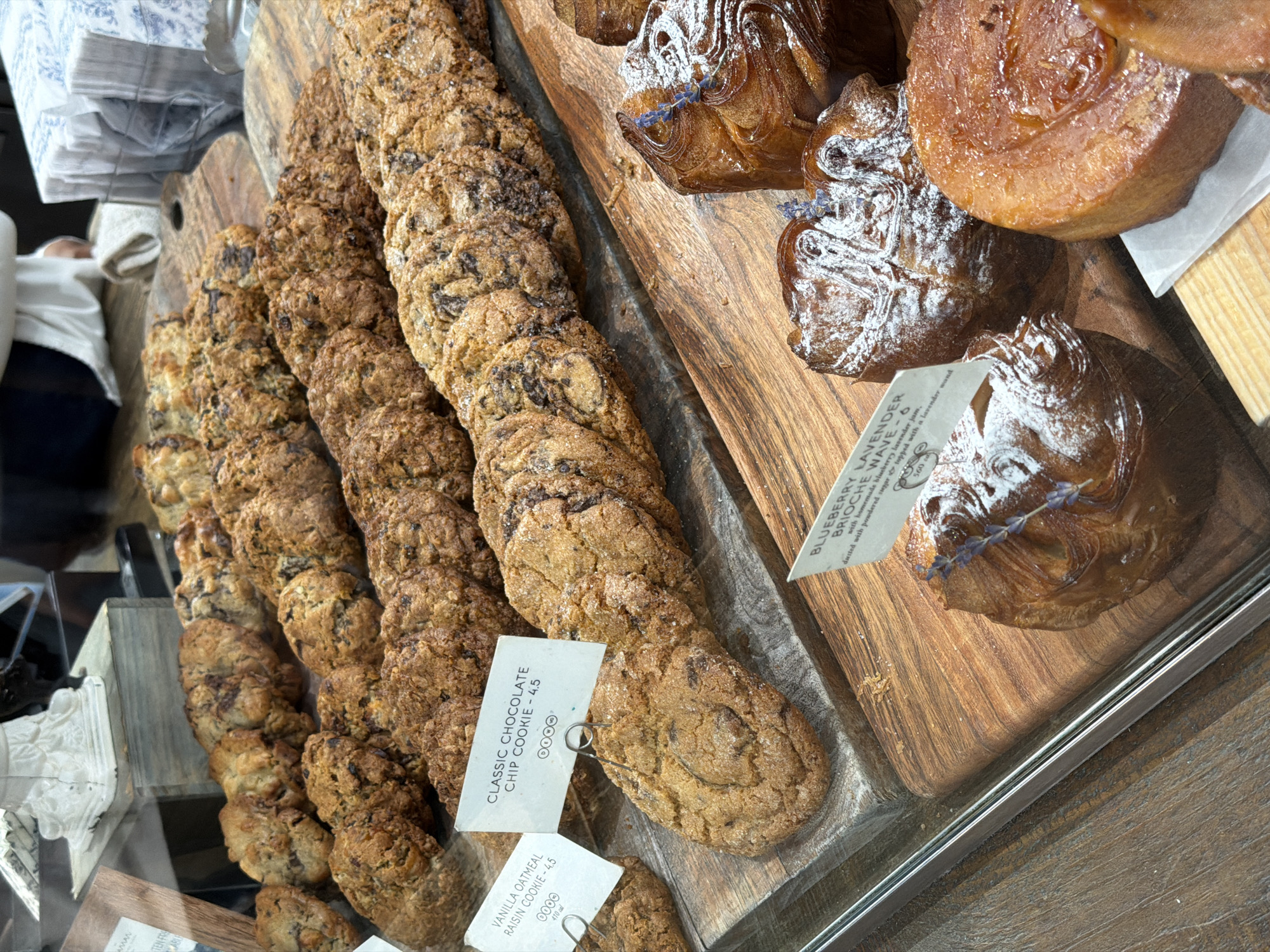This image captures a close-up view of a baked goods display inside what appears to be a bakery or cafe in New York, United States. A variety of cookies and pastries are neatly arranged on wooden boards within a glass display case, showcased for sale.
On the left side of the display, long rows of cookies are visible. The foreground prominently features "CLASSIC CHOCOLATE CHIP COOKIE - 4.5," with its tag clearly visible. Further back, another tag identifies "VANILLA OATMEAL RAISIN COOKIE - 4.5," also noting "410 cal." Both types of cookies appear golden brown with visible chocolate chips or raisins, respectively, and some have a sugar glaze or dusting.
On the right side, various pastries are displayed on a separate wooden board. These include glossy, golden-brown pastries, some of which are dusted with powdered sugar and adorned with small sprigs of lavender. One descriptive tag reads: "BLUEBERRY LAVENDER BRIOCHE WAVE - 6," followed by details "with homemade blueberry lavender jam, dusted with powdered sugar topped with a lavender sprig" and "560" (calories). Another round, glazed pastry is visible in the upper right.
In the blurred background on the far left, a person wearing a white shirt is partially visible, indicating a retail environment. The overall lighting suggests an indoor setting, highlighting the textures and colors of the baked goods.
