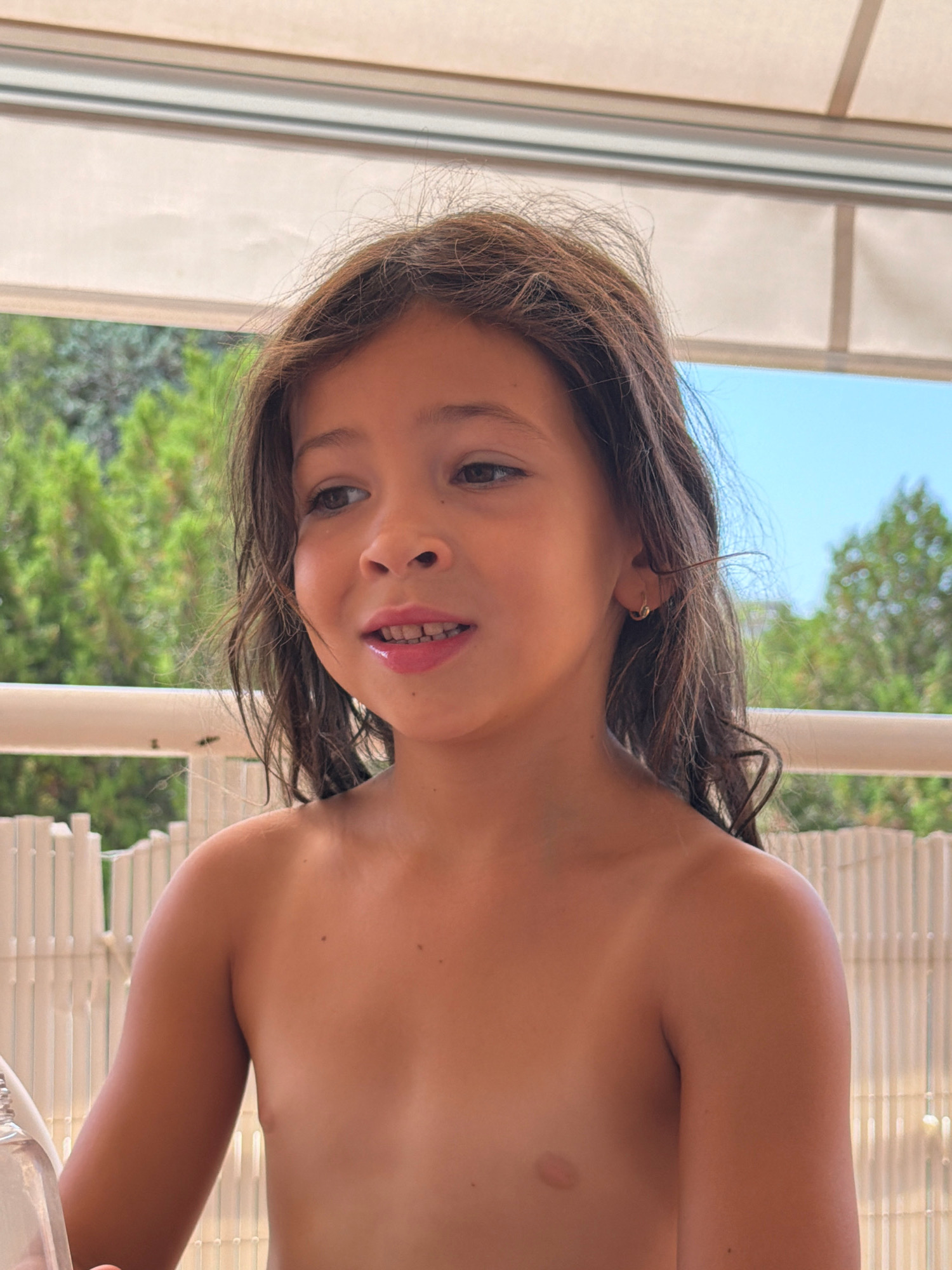 The image features a young girl, appearing to be around 5-7 years old, with long, dark brown hair and light brown eyes. She is looking to her left with a slight smile, her mouth slightly open. She is wearing a small gold hoop earring in her left ear. She is topless, with her shoulders and upper chest visible. Her skin is tanned.
The setting appears to be outdoors, likely on a balcony or patio, indicated by a white railing and a white fence-like structure in the background. Behind the fence, lush green trees and a bright blue sky are visible, suggesting a sunny day. The girl is positioned in front of a neutral-colored awning or roof structure.
The time of day is likely daytime, given the bright sunlight casting shadows and illuminating the scene. The overall mood of the image is relaxed and cheerful, with the girl's expression conveying a sense of lightheartedness.
There are no visible text elements in the image.
The location context provided is Salou, Spain, which aligns with the outdoor, sunny setting and the casual attire of the subject.