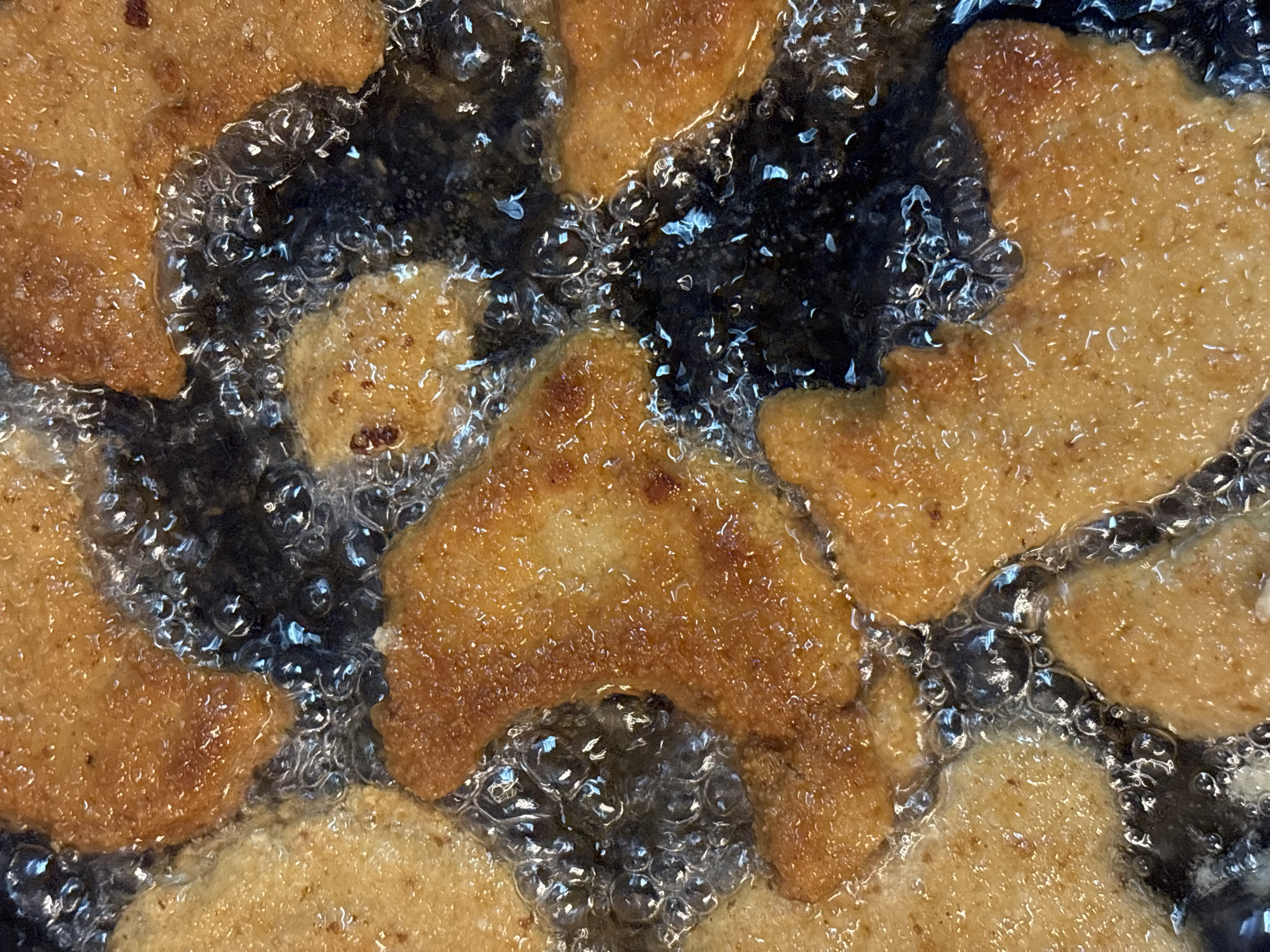 The image shows several breaded and fried food items cooking in oil. The items are golden brown and surrounded by bubbles of oil. The background is dark and indistinct due to the depth of the frying pan. No people or specific location is identifiable.
