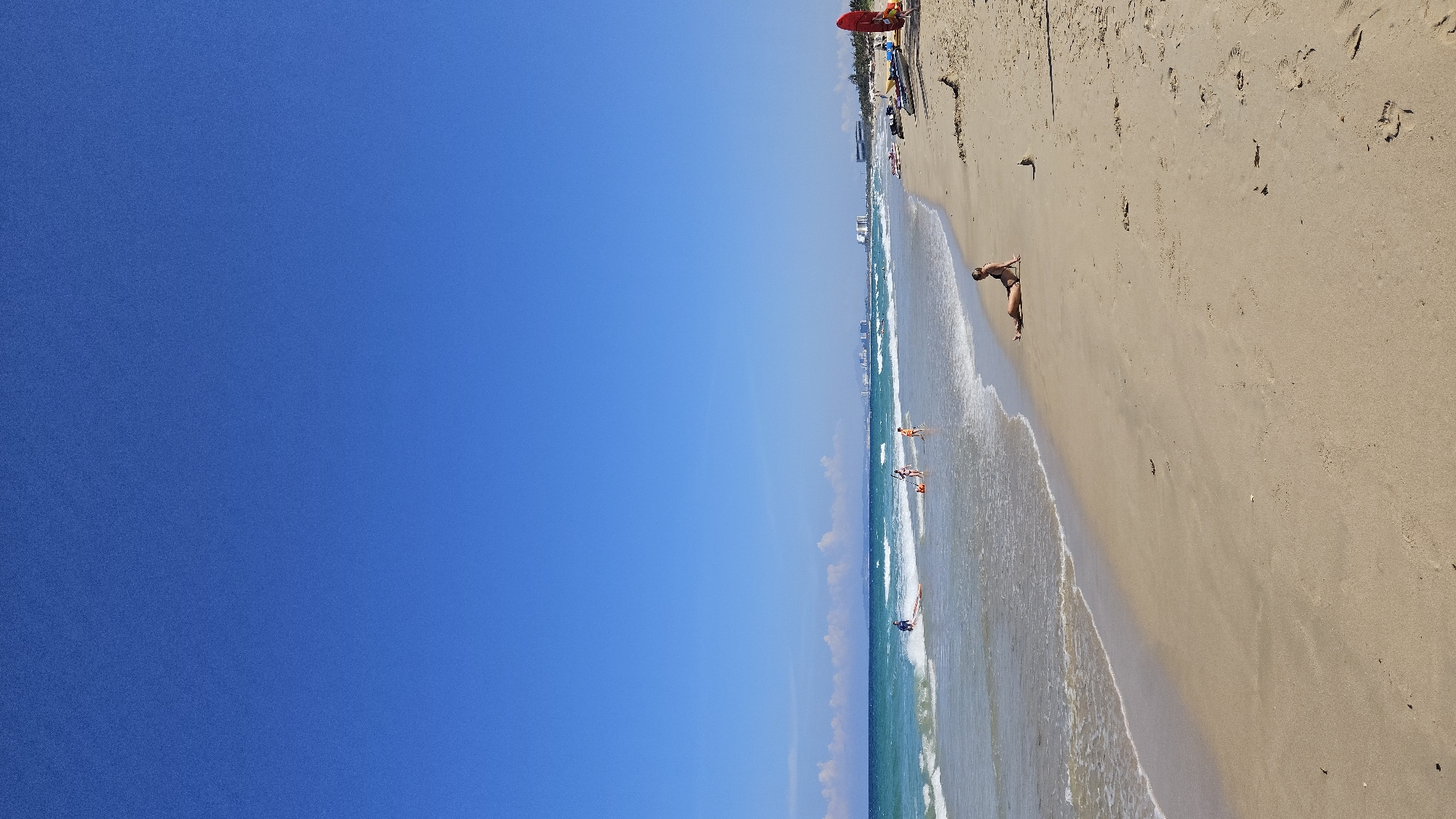 A horizontally-oriented, high-angle shot captures a sunlit beach scene.
The left half of the image shows a clear, blue sky.
The right half portrays a coastline. A sandy beach transitions into shallow, turquoise water with visible waves. Several small groups of people are in the water. One person is walking along the beach near the shoreline. Further up the beach, a few lifeguard stands with watercraft are observed. In the background, faint outlines of buildings suggest a city skyline, identifying the location as a beach in Cam Lam, Vietnam.