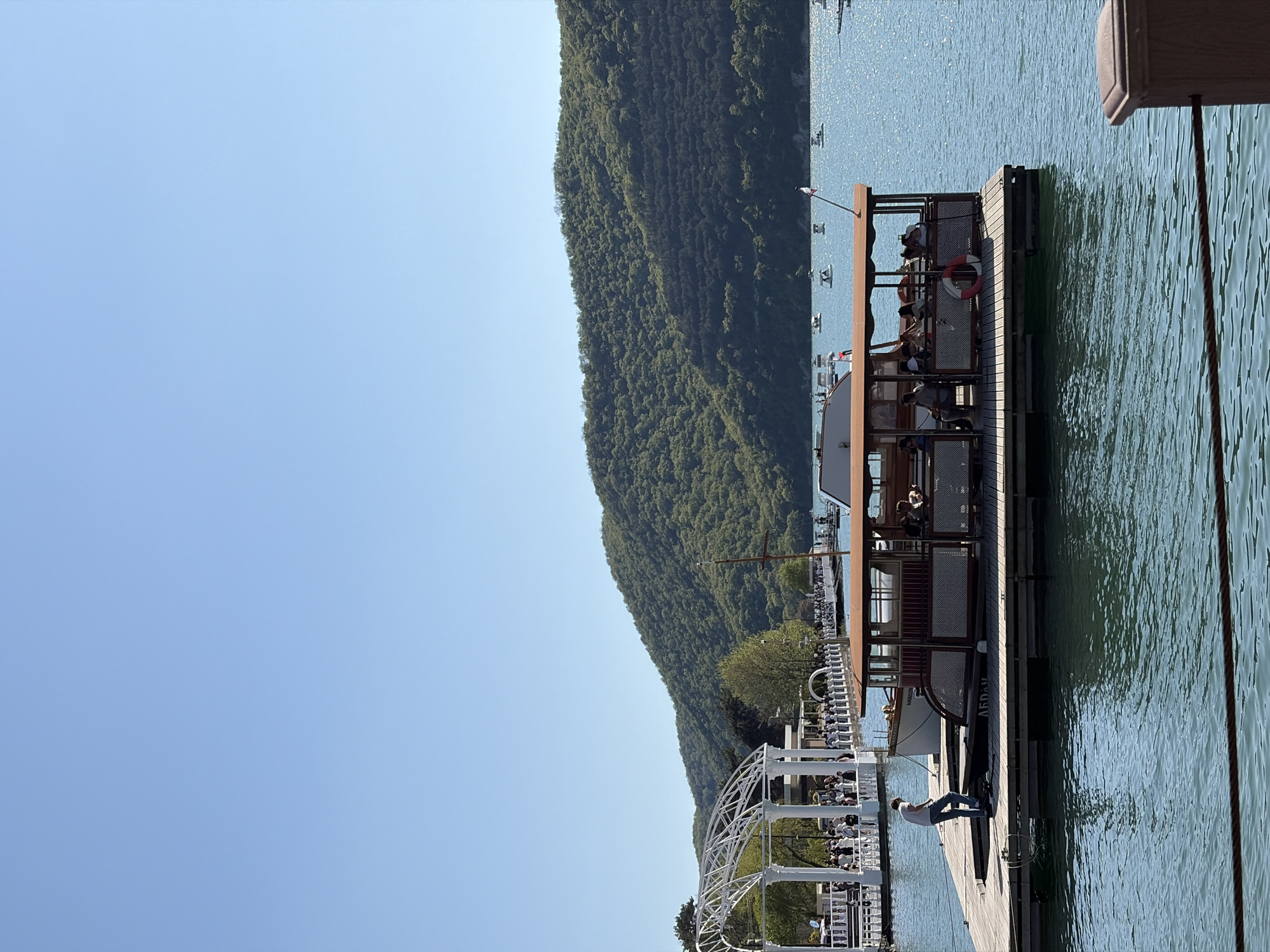 The image depicts a scene on a waterfront in Abrau-Dyurso, Russia. On a wooden pier stands a passenger boat or ferry. A man wearing a white shirt and blue jeans stands on the edge of the pier. The pier connects to a white decorative structure reminiscent of a gazebo. In the background, a steep, forested hillside rises. The sky is clear and blue. The water shows signs of slight ripples.