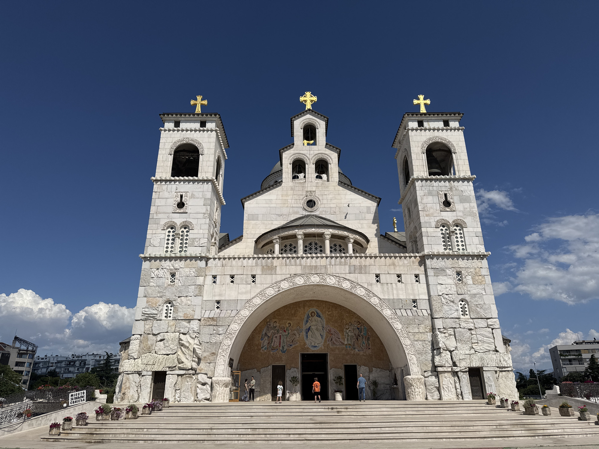 The image depicts the Cathedral of the Resurrection of Christ in Podgorica, Montenegro. It's a large white stone structure with three towers topped with golden crosses against a clear blue sky with some scattered clouds. The main entrance features a large archway with a mosaic above the doors. Several people are present on the steps leading to the entrance. There are flower pots lining the steps and surrounding the cathedral. Buildings are visible in the background, indicating an urban environment.