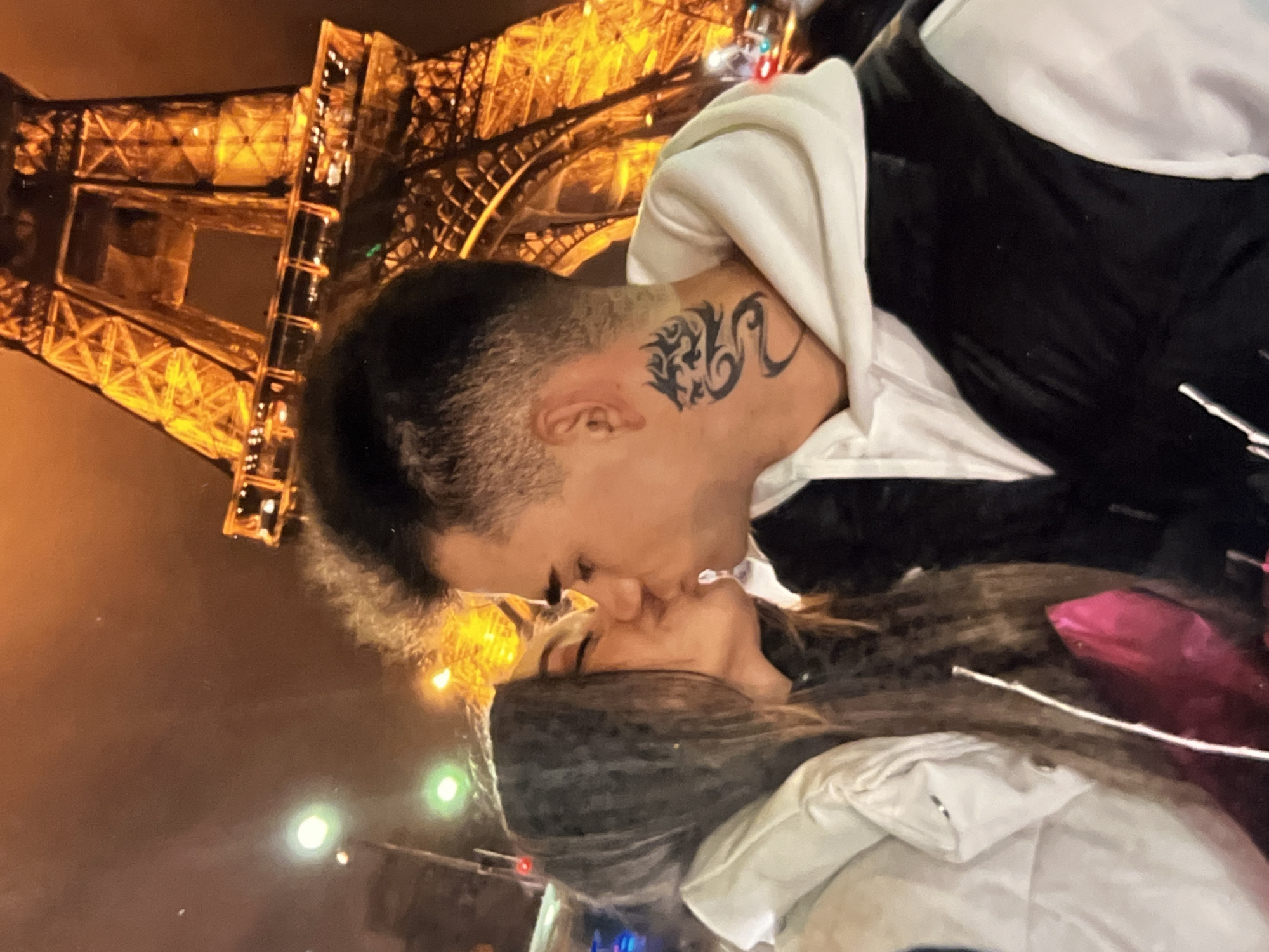 A couple is captured in a close-up shot, kissing in front of the illuminated Eiffel Tower at night. The woman, on the left, has long, dark, wavy hair and wears a white puffy jacket. The man, on the right, has short dark hair and a dragon tattoo on his neck. He is wearing a white hoodie underneath a black vest. The iconic Eiffel Tower looms in the background, its golden lights creating a warm glow against the dark sky. Streetlights and possibly other distant city lights are visible, suggesting an urban environment. The couple's interaction is intimate, focusing on their kiss, with the Eiffel Tower serving as a romantic backdrop. The scene is photographed from a slightly low angle, emphasizing the scale of the tower. There are no overt indications of violence or distress. The overall ambiance is romantic and celebratory.