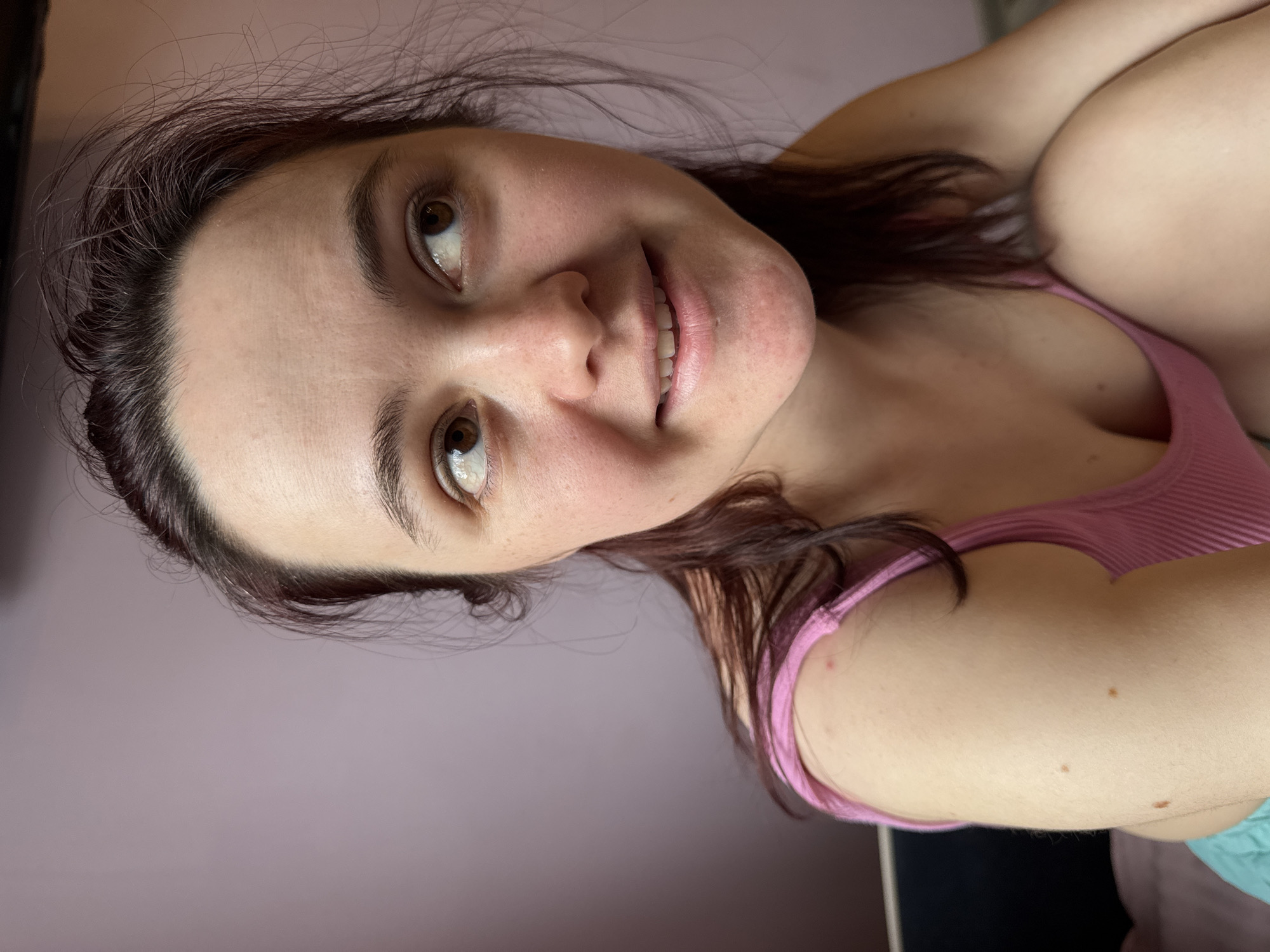 A woman with dark hair and brown eyes is smiling at the camera. She is wearing a pink tank top. The background is a light purple wall. The photo appears to have been taken indoors. There is no evidence to suggest the location is Sochi, Russia.