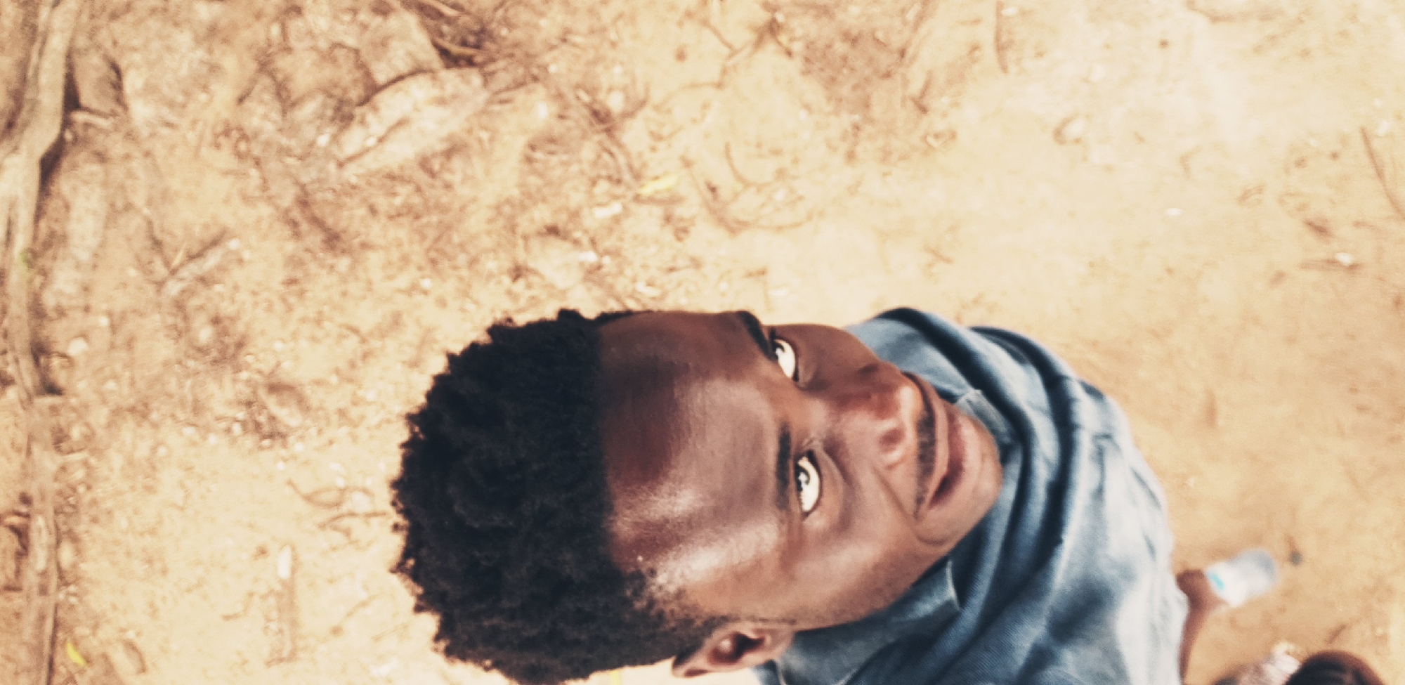 A vertical, eye-level, high-angle shot captures a dark-skinned man from the chest up, his head tilted upwards and to the right, with his gaze directed beyond the frame. He is wearing a blue collared shirt, which appears to be made of a soft material like cotton or denim. His short, dark, curly hair is neatly cut. The man's facial expression suggests he is engaged in conversation or looking at something of interest. He has a faint mustache and a subtle smile. The background consists of dry, sandy ground with scattered debris, possibly twigs or dried leaves. The lighting suggests it is daytime, with the sun casting subtle shadows. The overall scene appears to be outdoors, possibly in a dusty or unpaved area. Without specific landmarks or context, pinpointing the exact location within Narayi, Nigeria, is not possible based solely on the visual information provided.