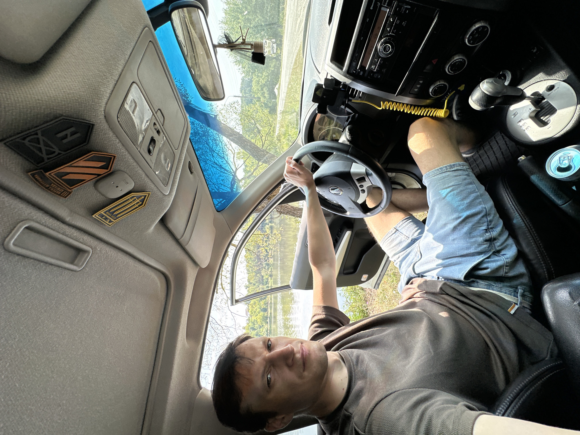 A man sits in the driver's seat of a car, which appears to be parked near a body of water with trees and foliage on the opposite bank. The car's interior is visible, including the steering wheel, dashboard, and gear shift. The man is looking towards the camera. On the ceiling of the car, several patches are affixed. The view from the car's window shows a scenic outdoor environment.