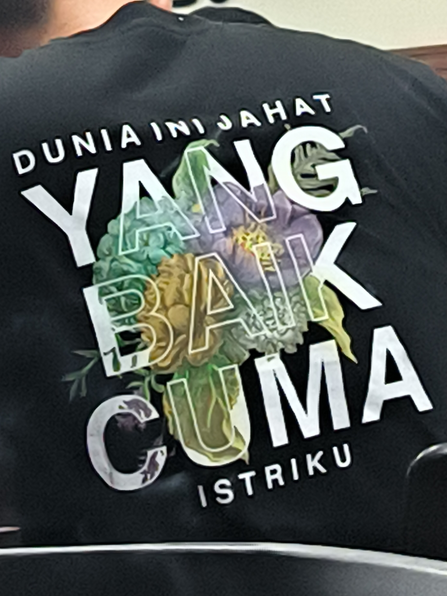 A close-up, rear-view shot features a person wearing a black t-shirt, likely in Lhokseumawe, Indonesia. The upper back and a portion of the person's head are visible, with the primary focus on the design printed on the t-shirt.
The black t-shirt displays a multi-line text design in white, along with a colorful floral graphic. The text reads:
"DUNIA INI JAHAT" (This world is evil/bad)
"YANG BAIK" (The good one)
"CUMA" (Only)
"ISTRIKU" (My wife)
The words "YANG BAIK CUMA" are significantly larger and appear somewhat translucent, revealing a detailed illustration of flowers and leaves beneath them. This floral graphic incorporates shades of green, purple, and yellow/orange, creating a vibrant contrast against the black fabric. The text "DUNIA INI JAHAT" and "ISTRIKU" are positioned above and below the larger, floral-overlaid text, respectively. The overall message conveys a humorous and affectionate sentiment about the wearer's wife being the sole good thing in a perceived 'evil' world. The background is out of focus, providing minimal details about the surroundings, but suggests an informal or casual setting. No specific activity is discernible other than the person wearing the T-shirt.