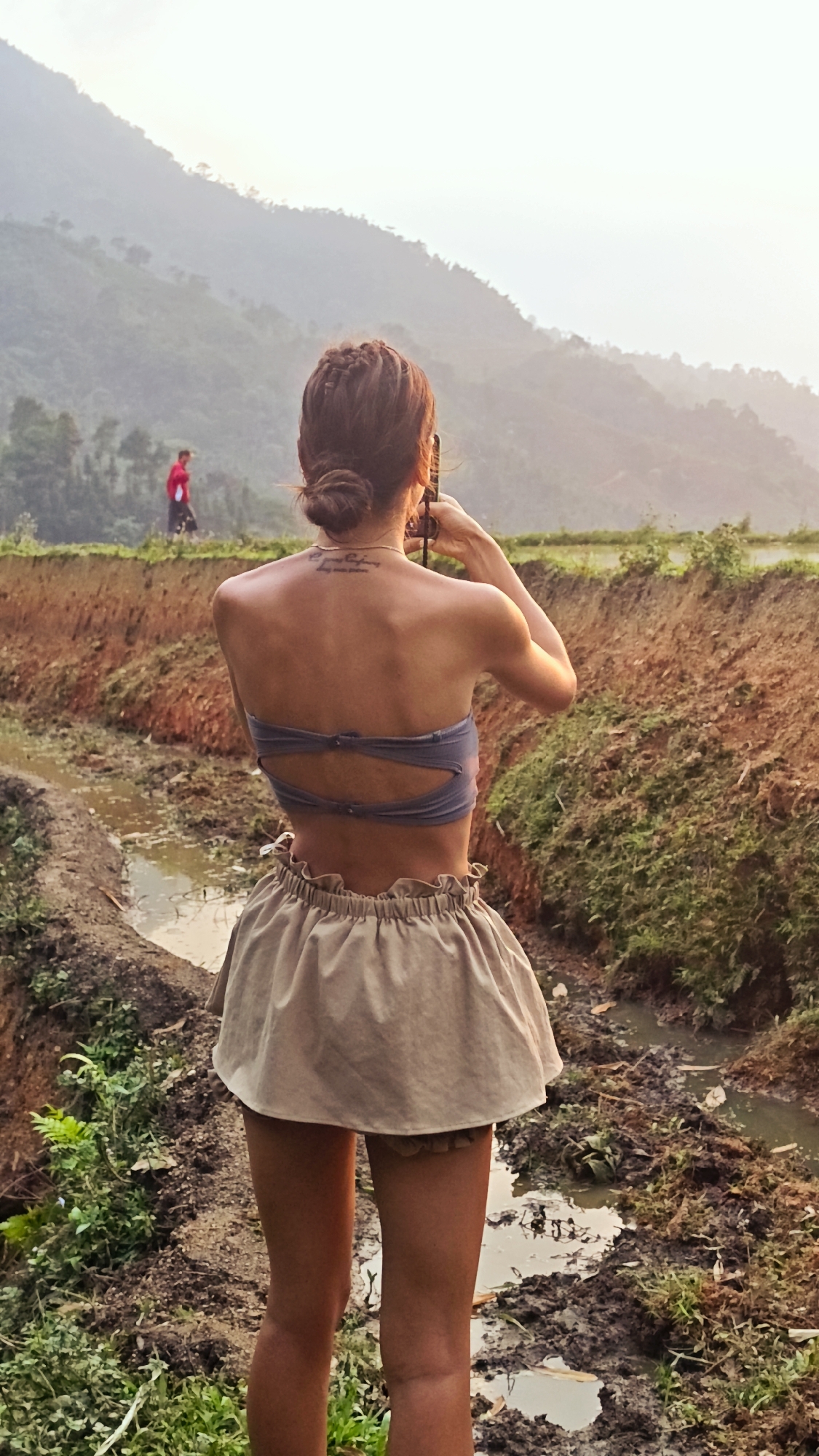A person, seen from behind, stands in a rural landscape in what appears to be Nậm Dịch Commune, Vietnam. The individual, with light brown hair styled in a bun, wears a grey strapless top with cut-out details on the back and a light beige, ruffled mini-skirt. They are holding a smartphone or small camera to their right eye, seemingly capturing the view.
The foreground and mid-ground consist of a muddy, water-filled channel or stream bordered by uneven earthen banks. These banks show patches of green vegetation mixed with exposed reddish-brown soil. In the distance, on a raised path to the left, another figure, possibly a man in a red shirt and dark pants, is walking. The background features layers of misty, green mountains and hills, characteristic of a terraced agricultural region.
The scene is illuminated by the soft, warm light of late afternoon or "golden hour," creating a gentle haze over the distant peaks. A tattoo with multiple lines of script is visible on the main subject's upper back, below the neck. The overall atmosphere suggests a serene and natural setting.