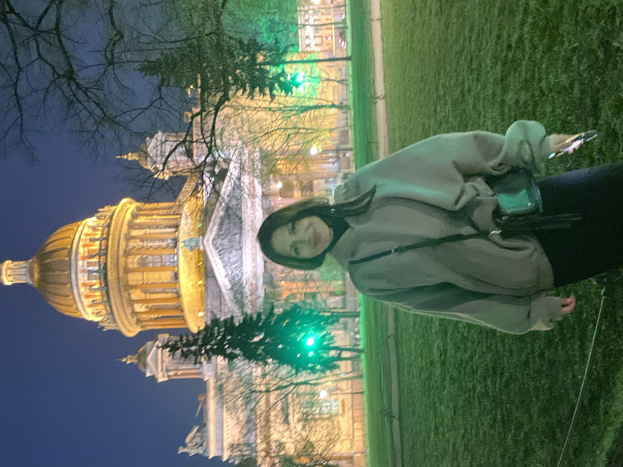 A medium shot presents a woman standing on a grassy lawn in front of Saint Isaac's Cathedral in Saint Petersburg, Russia. The cathedral, lit up at night, dominates the background. The woman is wearing a grey hoodie and black pants. She holds a phone in her right hand. Her left hand is at her side. The scene is composed with a blend of natural and artificial light. Bare tree branches are visible above the cathedral.
