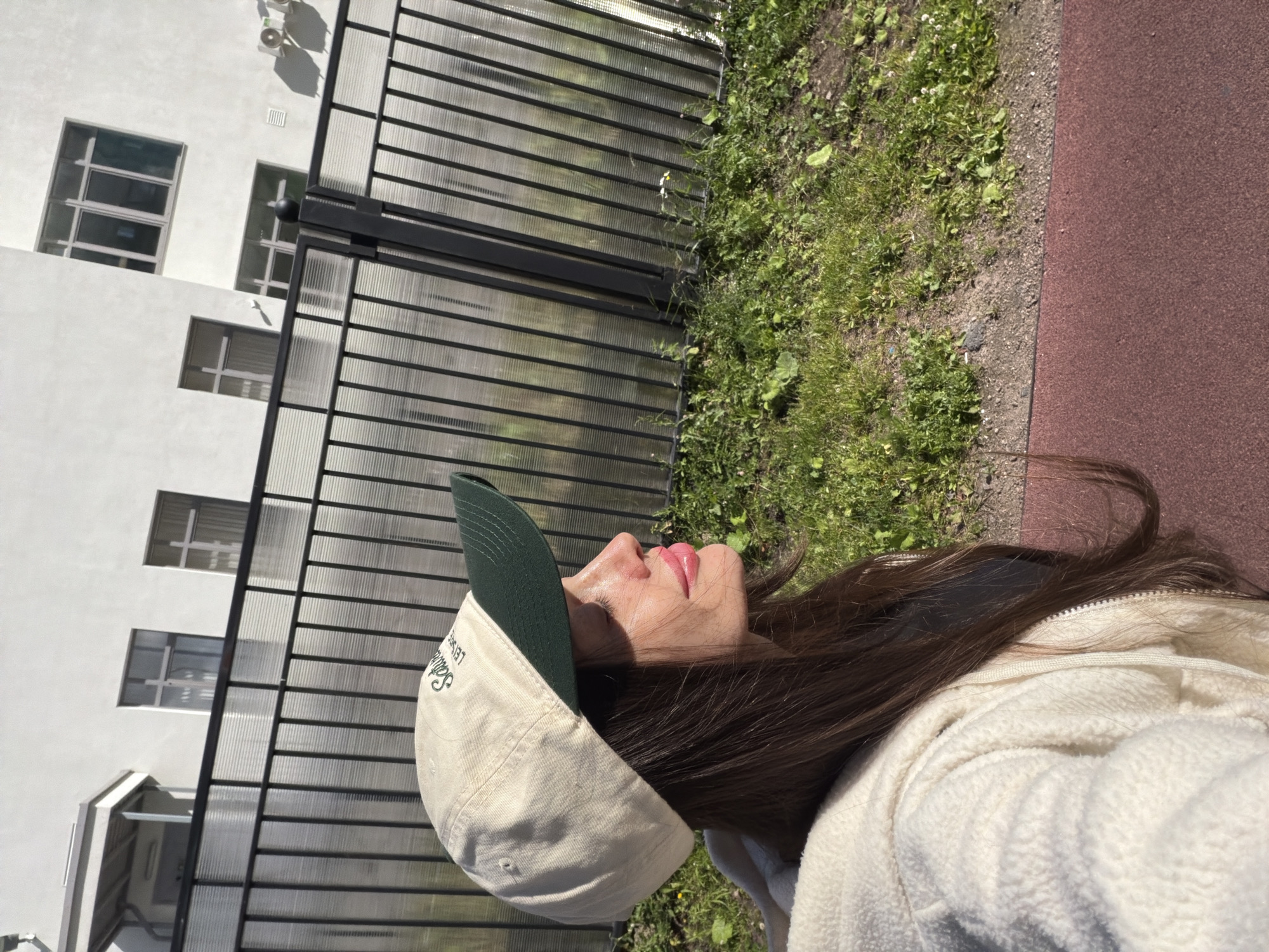 The media file shows a close-up of a person wearing a light-colored baseball cap with a dark green brim. The cap has embroidered text in green. The person's face is partially visible, with their eyes closed and lips slightly parted. They appear to be looking upwards, with sunlight on their face. The person has long, dark brown hair that is partially visible, and is wearing a cream-colored textured jacket.
The background suggests an outdoor setting. To the left, there is a white building with multiple windows. In the middle ground, there is a black metal fence with translucent panels, behind which is a grassy area with some scattered weeds and dirt. To the right, there is a reddish-brown paved surface, possibly a path or sports track.
The lighting indicates it is daytime, likely sunny, given the strong shadows and highlights on the person's face and the building. The overall scene gives the impression of a casual outdoor moment, possibly in a park or near a residential area, given the building and fence. The person's expression could suggest relaxation or enjoyment of the sun.
The visible text on the cap appears to be "SALLAT LEISURE".