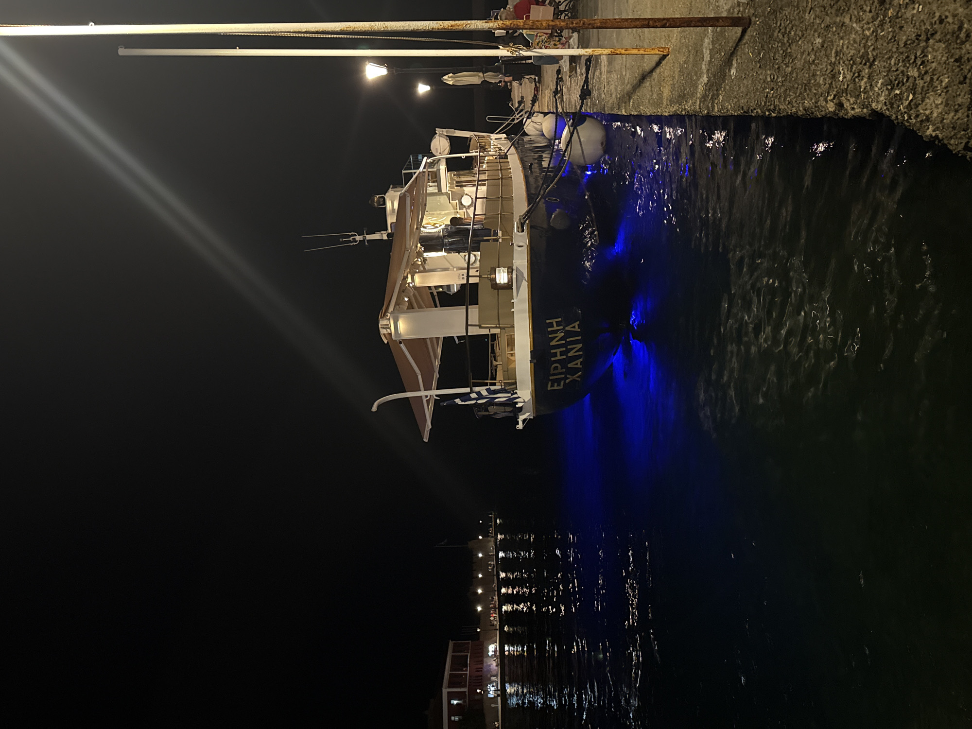 A night view captures a boat docked at a harbor. The boat, with "EIPHNH XANIA" inscribed on its side, is illuminated by blue lights underneath its hull. A Greek flag is attached to the boat's railing. In the background, a red building with illuminated windows stands across the water, alongside a row of streetlights and a fortified wall. The water reflects the lights from the building and the boat. The foreground shows a textured concrete dock with two vertical poles and some seating. The scene is lit by artificial lights, creating a contrast between the dark night sky and the illuminated elements.