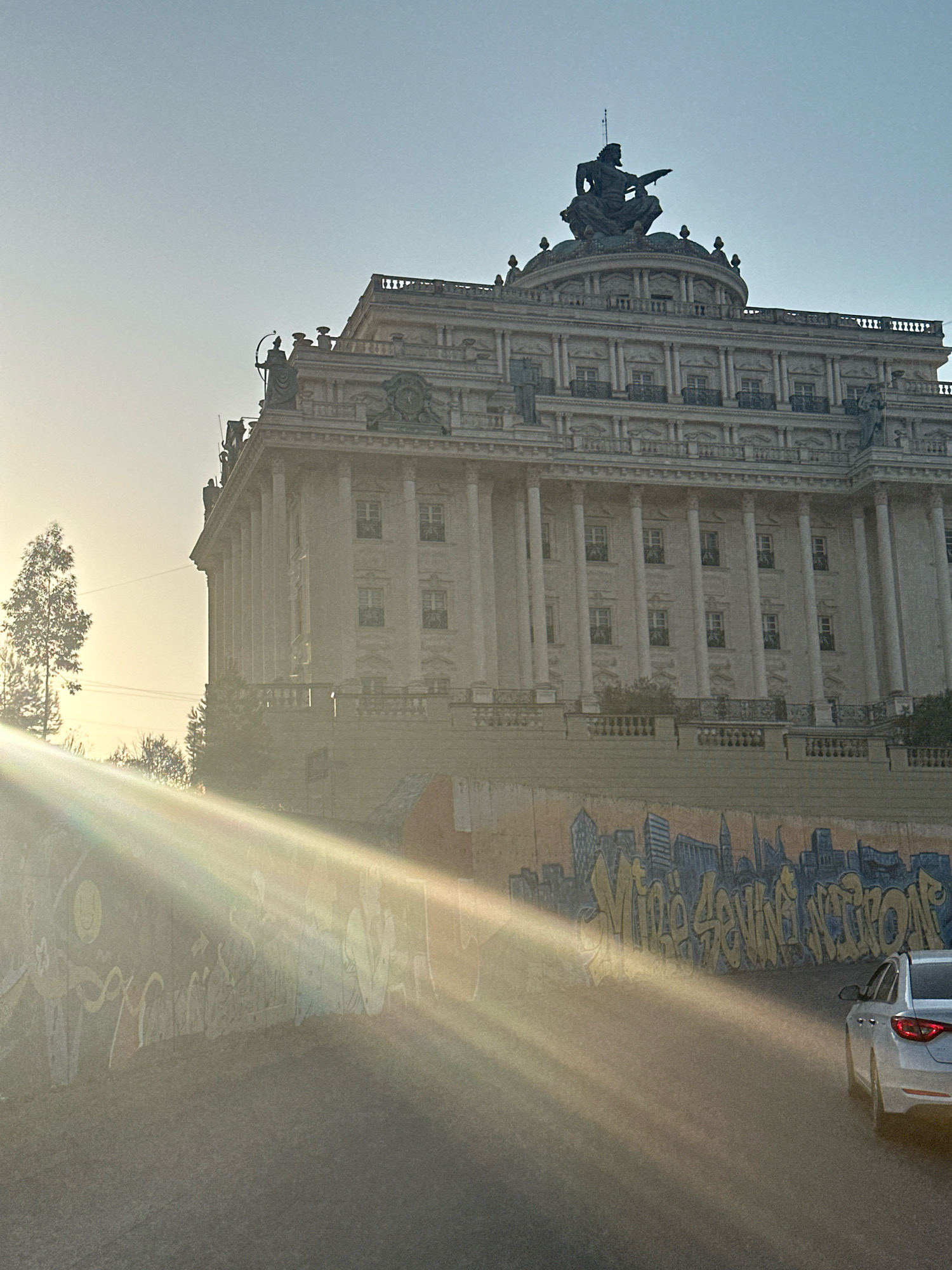 The image depicts a grand, classical-style building with a large statue on its roof. The building is made of light-colored stone and features numerous columns and windows. In the foreground, a mural covers a wall, with graffiti tags and city skyline imagery. Sunlight streams across the scene, creating lens flare effects and casting shadows. A white car is partially visible on the right side of the image. The sky is a pale blue with a gradient towards a lighter hue at the horizon. A single tree stands to the left of the building.