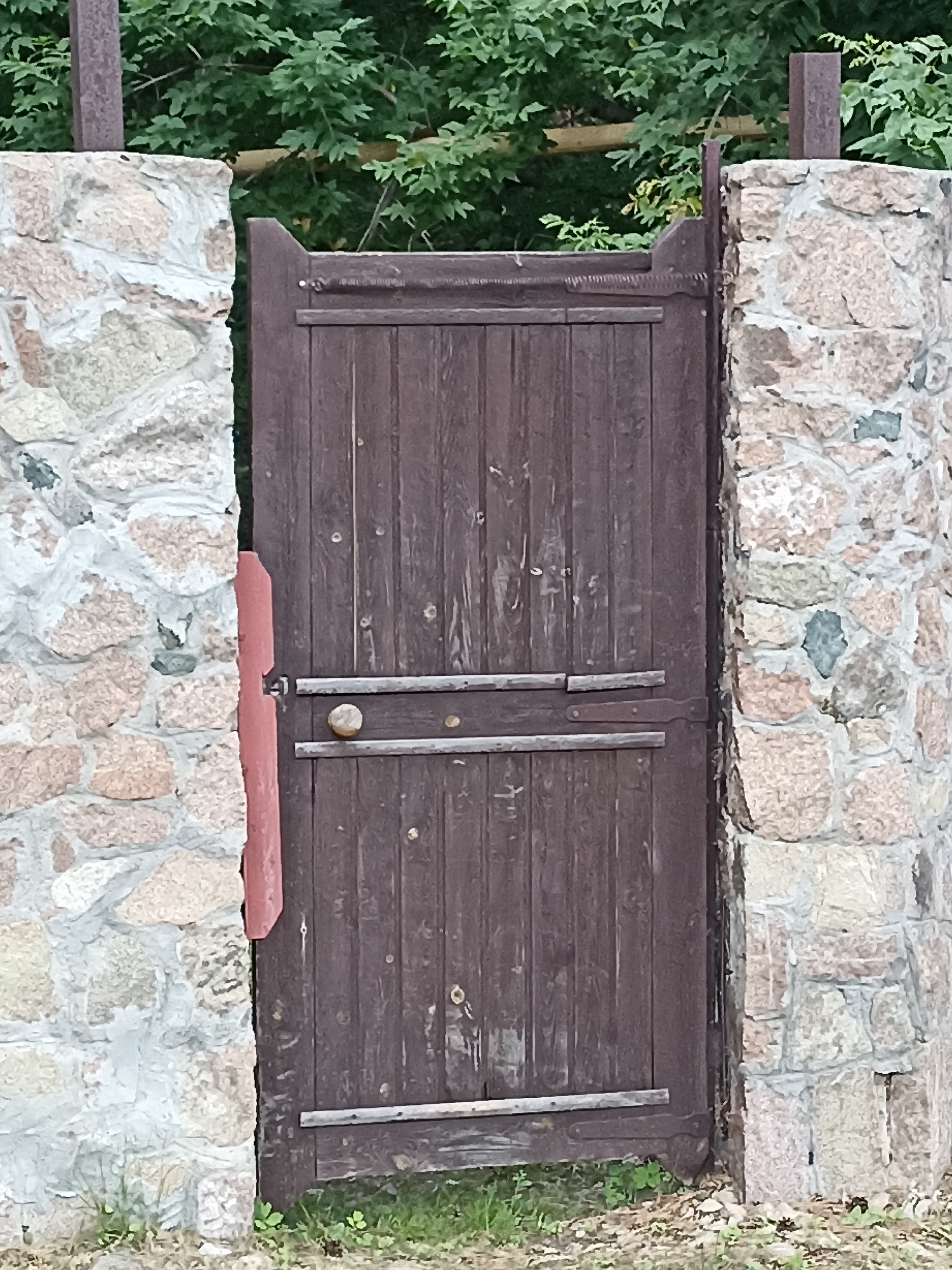 A wooden gate, painted dark brown, is set between two stone pillars. The gate is made of vertical wooden planks and has two horizontal metal bars across its front for support. A circular metal knob serves as a handle. The stone pillars are constructed with rough-hewn, light-colored stones of varying sizes, mortared together. Behind the gate and pillars, dense green foliage, presumably trees or bushes, is visible. The ground in front of the gate is covered with dry leaves and sparse green grass. The scene appears to be an outdoor entrance to a property. There are no people visible in the image. This image is a close-up shot of the gate and pillars, with the background foliage providing context.
