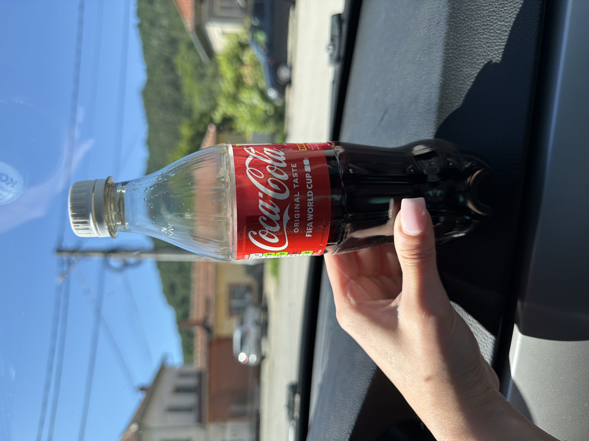 A partially full Coca-Cola bottle is held by a person's left hand, with their fingers wrapped around the lower portion of the bottle. The bottle has a red label with the Coca-Cola logo and text that reads "ORIGINAL TASTE FIFA WORLD CUP". The bottle is positioned in front of a car's dashboard, casting a shadow. The background, viewed through what appears to be a car window, shows a sunny outdoor scene with houses, trees, and a clear blue sky with some utility lines. The location is identified as Beli Osam, Bulgaria.
