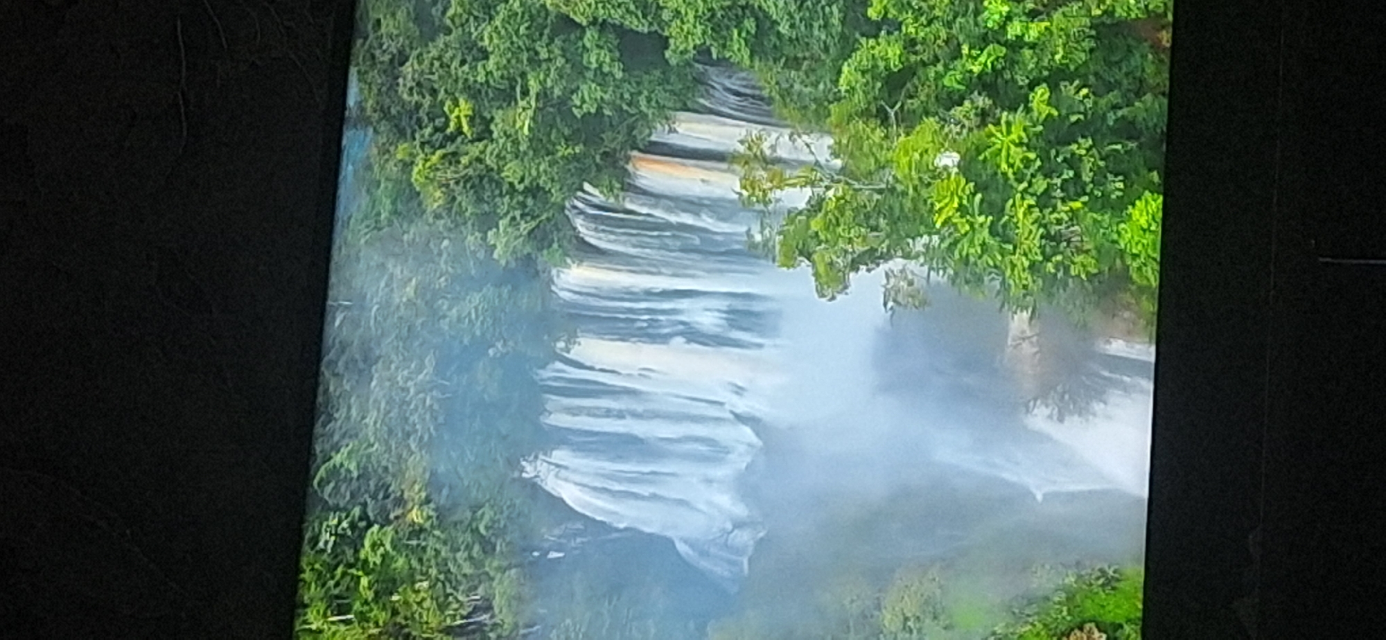 The image shows a wide waterfall cascading down a rocky cliff face, surrounded by lush green foliage. The water is white and frothy, creating a mist at the base of the falls. The surrounding environment appears to be a natural landscape, possibly a rainforest or dense forest, with trees and vegetation covering the cliffs and slopes. The lighting suggests it may be daytime, with the sun illuminating the scene and highlighting the white water of the falls. The image does not provide any specific details or context that would place it in Bila Tserkva, Ukraine.