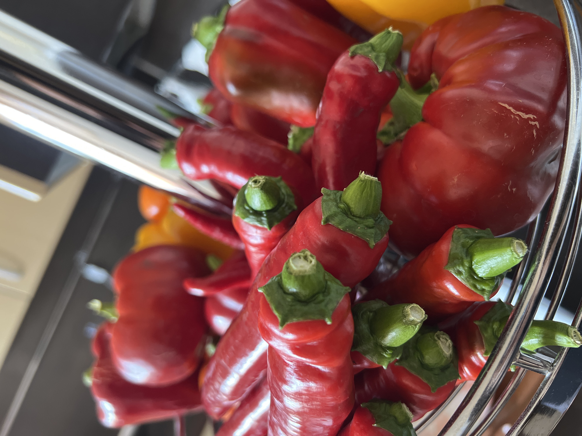 The image depicts a close-up, high-angle view of a metal basket filled with red bell peppers and red chili peppers. The peppers are vibrant in color and appear fresh. The basket is made of shiny, reflective metal and has a wireframe design. In the background, out of focus, are hints of a kitchen environment with what appears to be cabinetry and possibly a countertop. There are no people or animals visible in the image. The scene is indoors and appears to be in a domestic setting, likely a kitchen, but no specific location in Tolyatti, Russia can be identified from the visual information provided.
