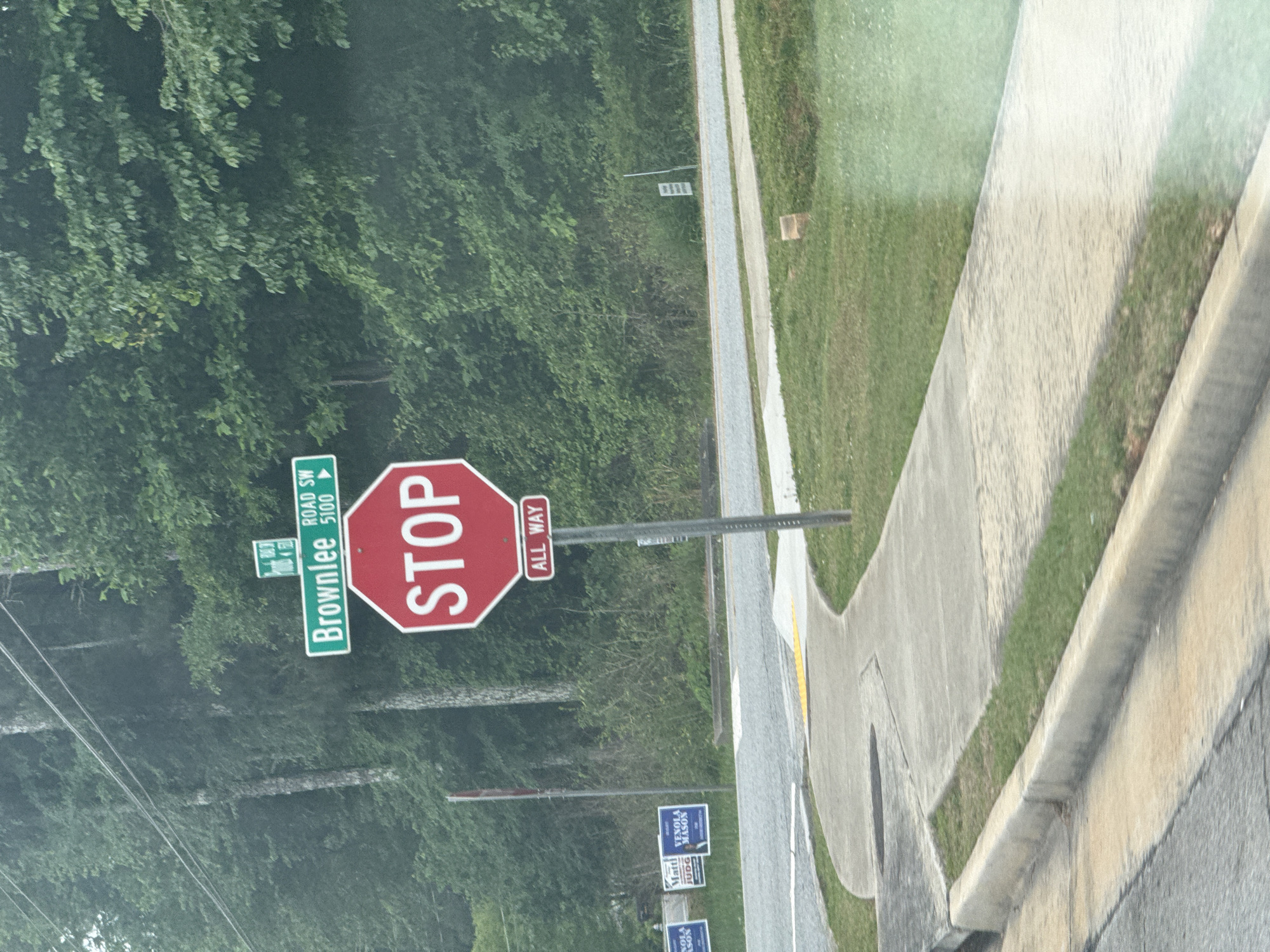 The image shows a street scene in Mountain Park, Georgia, United States. A "Brownlee Road SW 5100" street sign and an "All Way" stop sign are prominently featured. Political campaign signs for "Matt & Venola Mason Judge" are visible alongside the road. The scene includes a sidewalk and greenery with trees in the background. The shot appears to be taken from inside a vehicle.