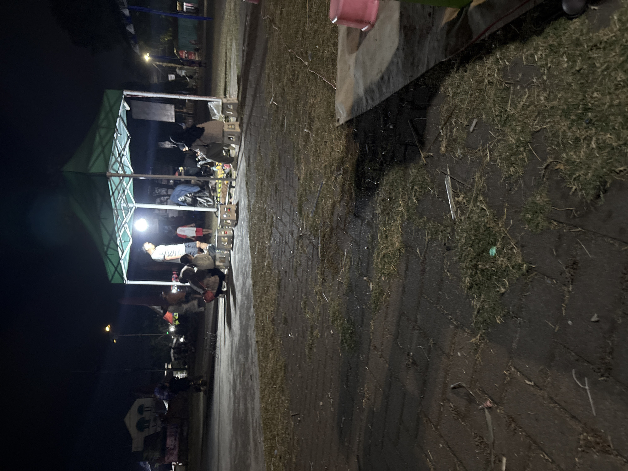 The image depicts a nighttime scene in Palimanan, Indonesia, possibly a marketplace or a gathering spot. A green canopy tent is erected, illuminated by a bright light, creating a central focus. Several people are gathered around and under the tent.
In the foreground, the ground is a mix of paved brickwork and patches of grass. Debris, such as small sticks and leaves, is scattered across the ground.
Under the tent, a man in a light-colored t-shirt and shorts stands, appearing to be interacting with others. Several individuals are seated on small, white stools. Some are wearing hijabs. There are also some objects that appear to be goods or equipment arranged around the tent. In the background, streetlights are visible, and there are indistinct structures, possibly buildings and other stalls, suggesting an urban or semi-urban environment. The overall atmosphere is casual and communal.