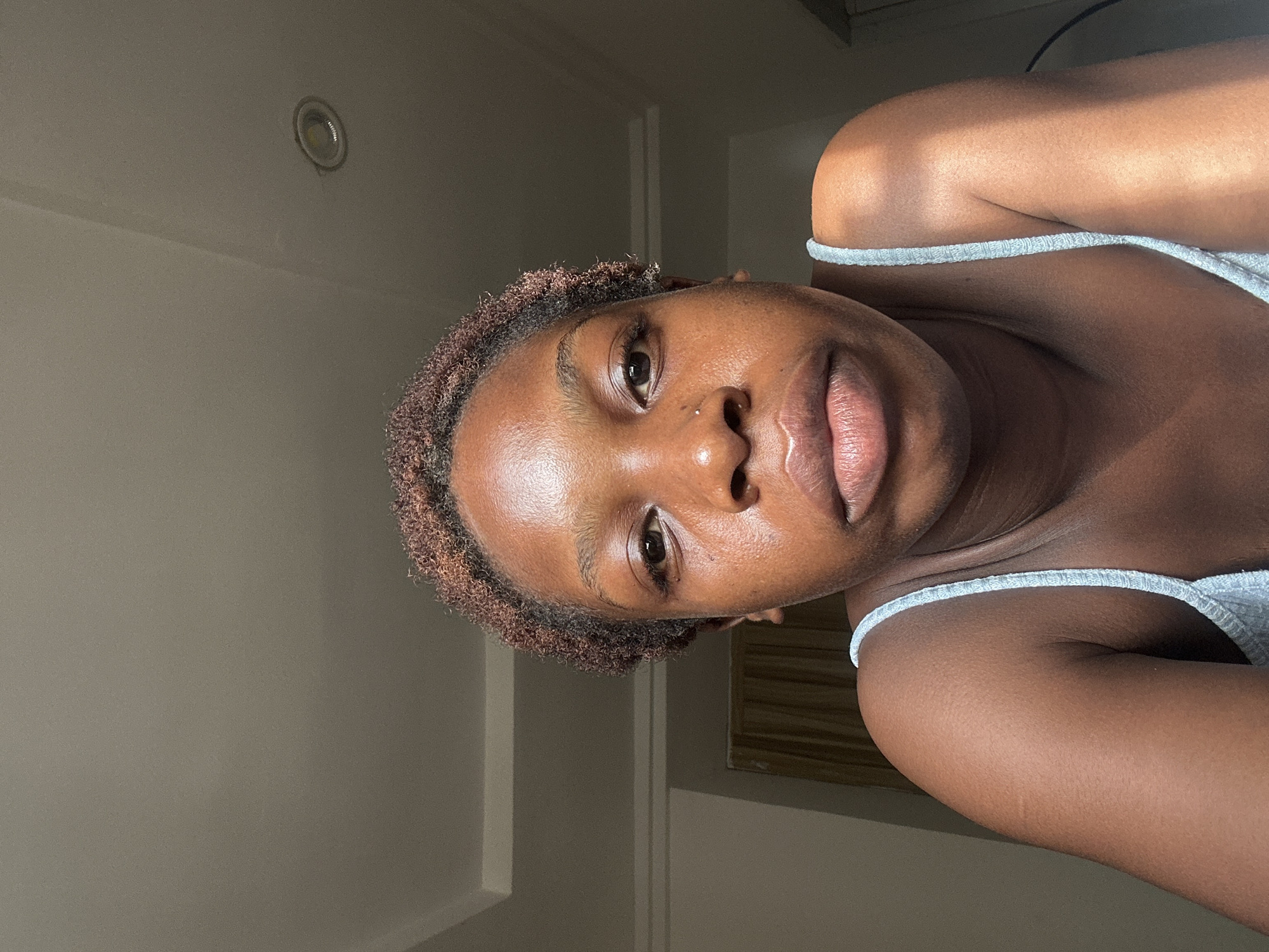 A selfie of a dark-skinned woman with short, brown hair. She is wearing a grey tank top. The background is a plain, light grey wall with a recessed light fixture. The location is likely indoors. There is no information to confirm the city of Abuja, Nigeria.