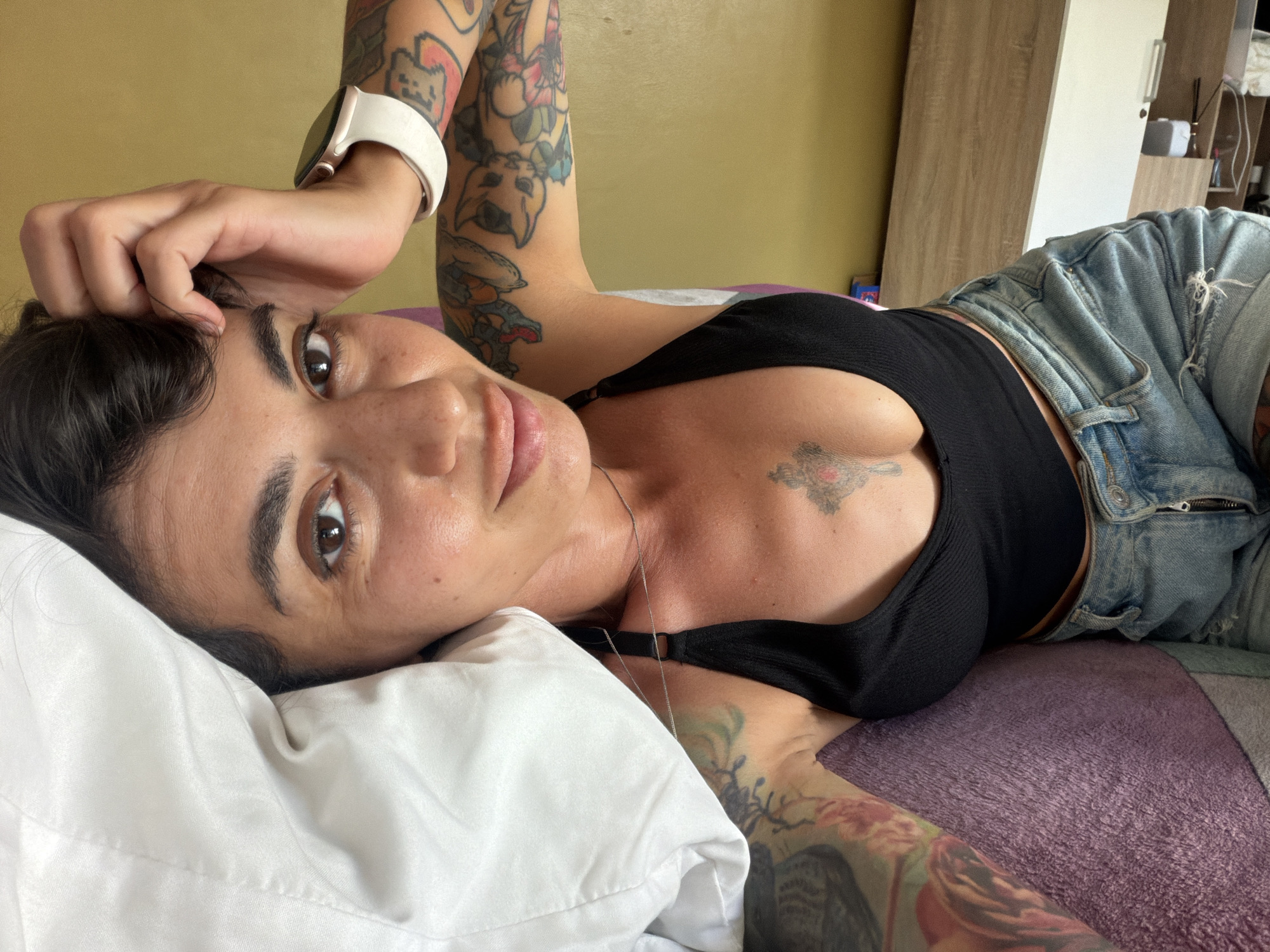 A woman with dark hair and tattoos lies on a bed with a white pillow. She is wearing a black tank top and denim shorts. Her left arm is raised, resting on her head, and she is wearing a white smartwatch. The room appears to be a bedroom, with a wooden wardrobe in the background. The lighting suggests it is daytime. The woman is looking towards the camera with a neutral expression.