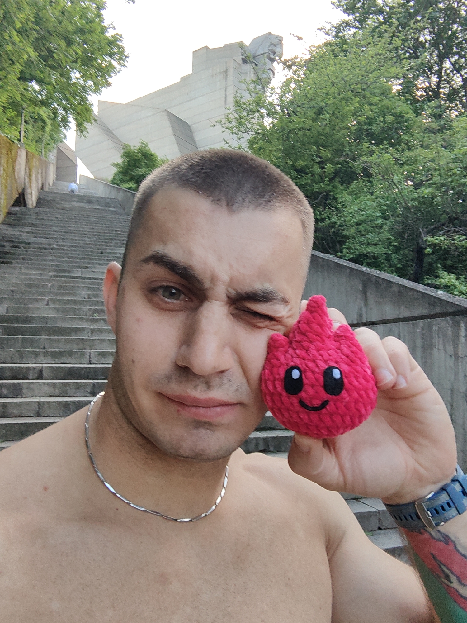 A shirtless man with a buzzcut stands on a set of concrete steps with metal railings in Shumen, Bulgaria. He is holding a small, red, heart-shaped plush toy with a stitched-on smile. The man is wearing a silver chain necklace and a blue sports watch. He is winking at the camera. Behind him, at the top of the steps, is the Monument to 1300 Years of Bulgaria. Trees flank either side of the steps. A person can be seen on the steps in the background. The overall environment suggests an outdoor setting on a clear day.