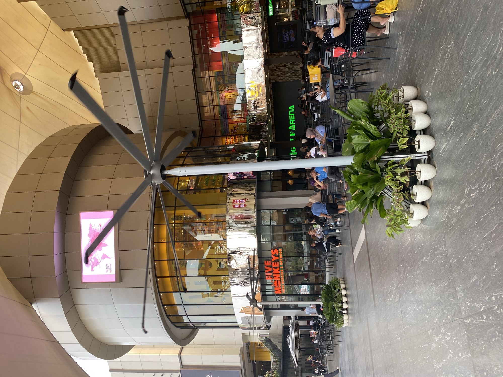 The image depicts an outdoor seating area of a commercial establishment, likely a mall or food court, situated in North Jakarta, Indonesia. The foreground is dominated by a large, industrial-style fan with numerous blades, positioned centrally and angled towards the viewer. Surrounding this fan are several potted plants arranged in a line. The midground reveals an array of black tables and chairs, occupied by individuals and groups of people seated and interacting. Some patrons are engaged in conversation, while others appear to be dining or relaxing. In the background, a multi-story building features a curved facade with large windows, behind which various shops and restaurants are visible. One prominent sign reads "FIVE MONKEYS FRESH BURGERS & HOTDOGS". Another sign, illuminated with green text, displays "IGLF ARENA". The overall environment suggests a bustling public space with commercial activity and casual dining. The architectural style of the building and the presence of the large fan point to a modern, possibly open-air or well-ventilated complex. The lighting indicates it is daytime, with a mix of natural and artificial light.