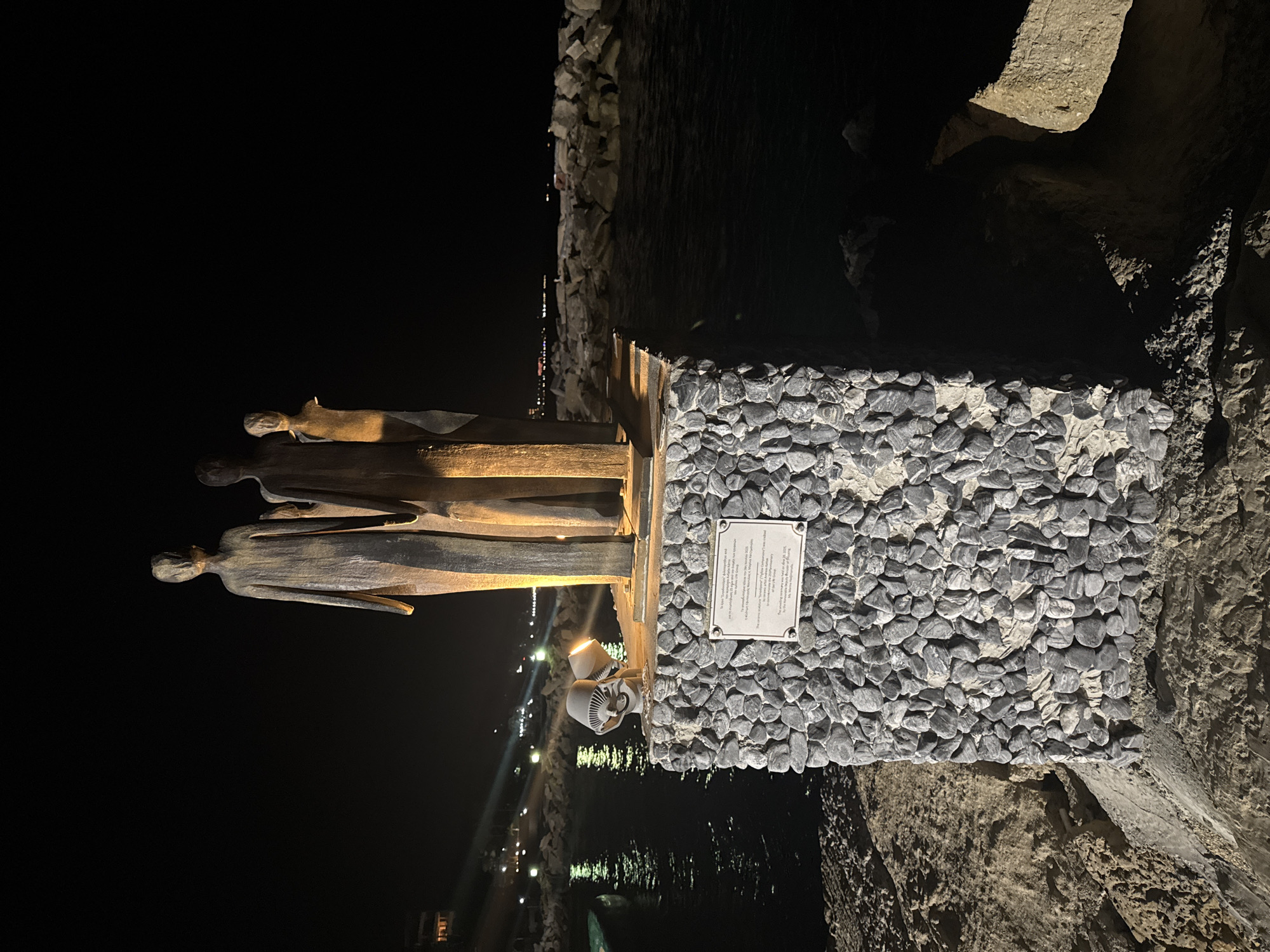 This is a nighttime outdoor shot of a sculpture on a stone pedestal near the sea in the city of Agios Tychona Community, Cyprus. The sculpture depicts three abstract human figures, two taller and one shorter, made of a dark material. The pedestal is constructed from small, gray, rounded stones. A plaque with text in Greek and English is affixed to the front of the pedestal. Two uplights, one angled towards the sculpture and the other illuminating the sea, are positioned on the pedestal. The sea is dark and appears choppy. Beyond the sea, a rocky breakwater stretches into the distance, with lights from a coastal town visible on the far horizon. The foreground is composed of sand and rocks.