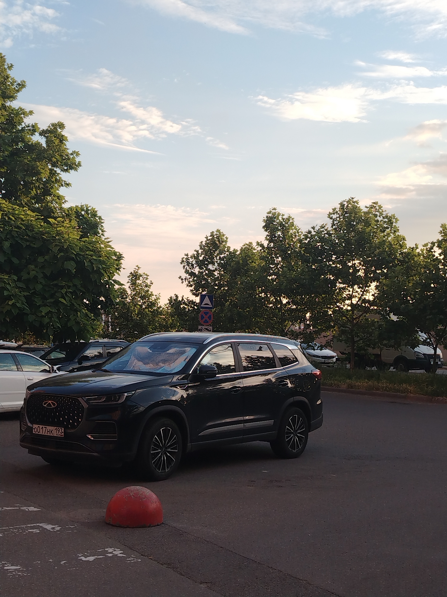 The image shows a black Chery Tiggo 7 Pro parked on an asphalt road in Krasnodar, Russia. The license plate of the vehicle is visible, reading "0017HK 193". A red concrete bollard is in the foreground. In the background, there are several other vehicles and lush green trees lining the street under a partially cloudy sky. The scene appears to be a typical urban streetscape.