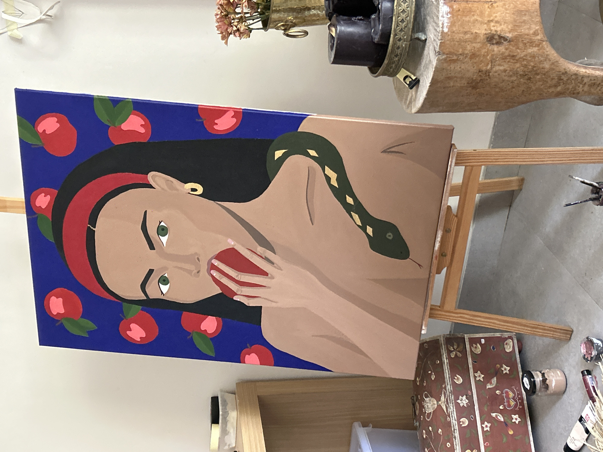 A painting of a woman with a snake is displayed on a wooden easel indoors. The woman is depicted with tan skin, dark hair with a red headband, and green eyes. She is eating a red apple. A green snake with yellow markings is draped over her shoulder. The background of the painting is a royal blue color with red apples scattered around. The painting sits on a wooden easel next to a small wooden table with an old-fashioned metal lamp on it. A painted wooden chest sits below the easel, and paint supplies are scattered on the floor nearby. The location is likely an art studio or home in North Kuta, Indonesia.