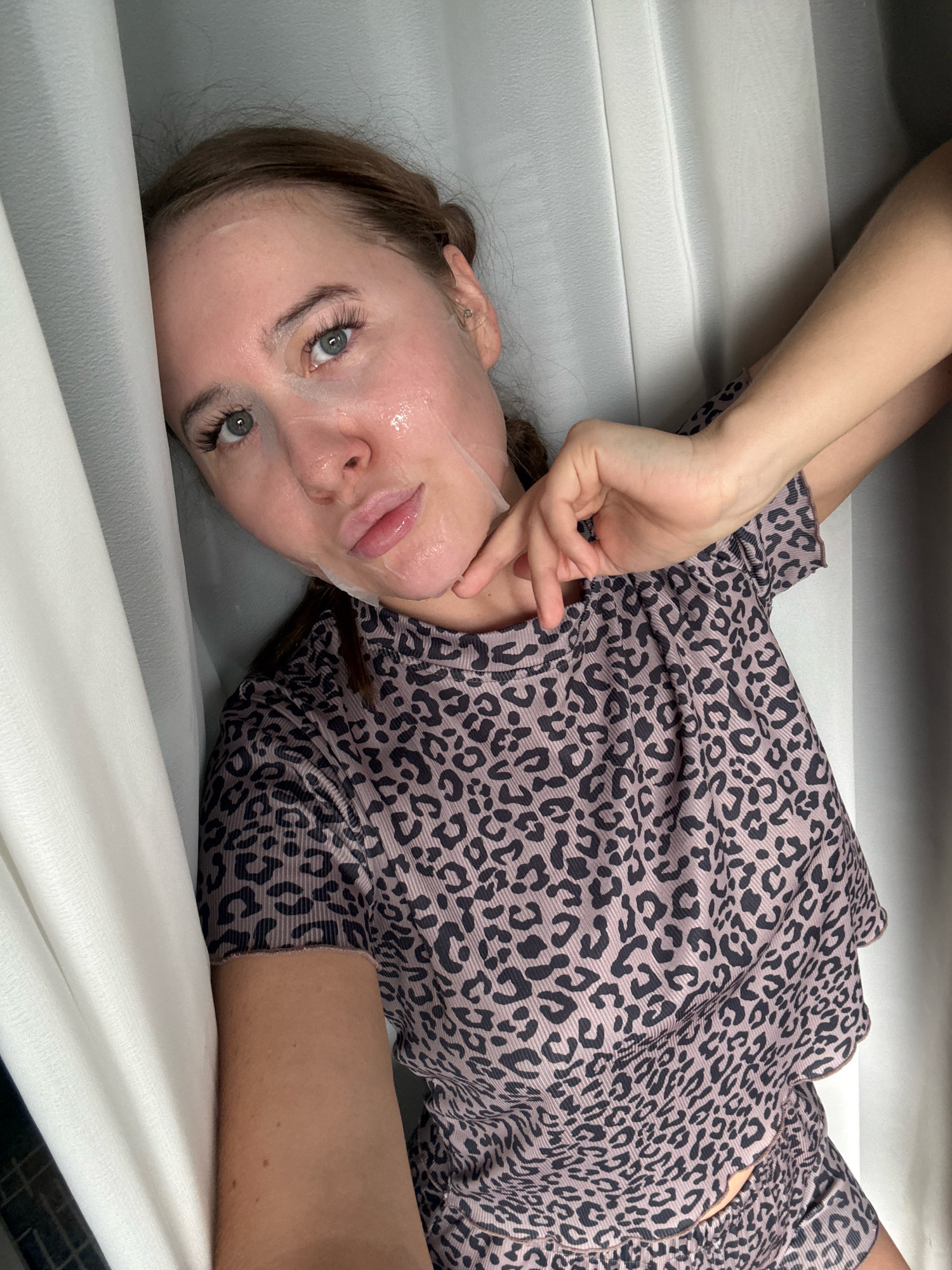 A young woman with light skin and brown hair tied back is leaning against a white curtain, her head tilted to the left. She is wearing a leopard-print shirt and has a sheet mask on her face. Her right hand is raised, with her fingers near her chin. The background consists of white curtains, creating a soft, diffused light. The setting does not provide any specific information about the city of Yiwu, China, as it is a generic indoor environment.
