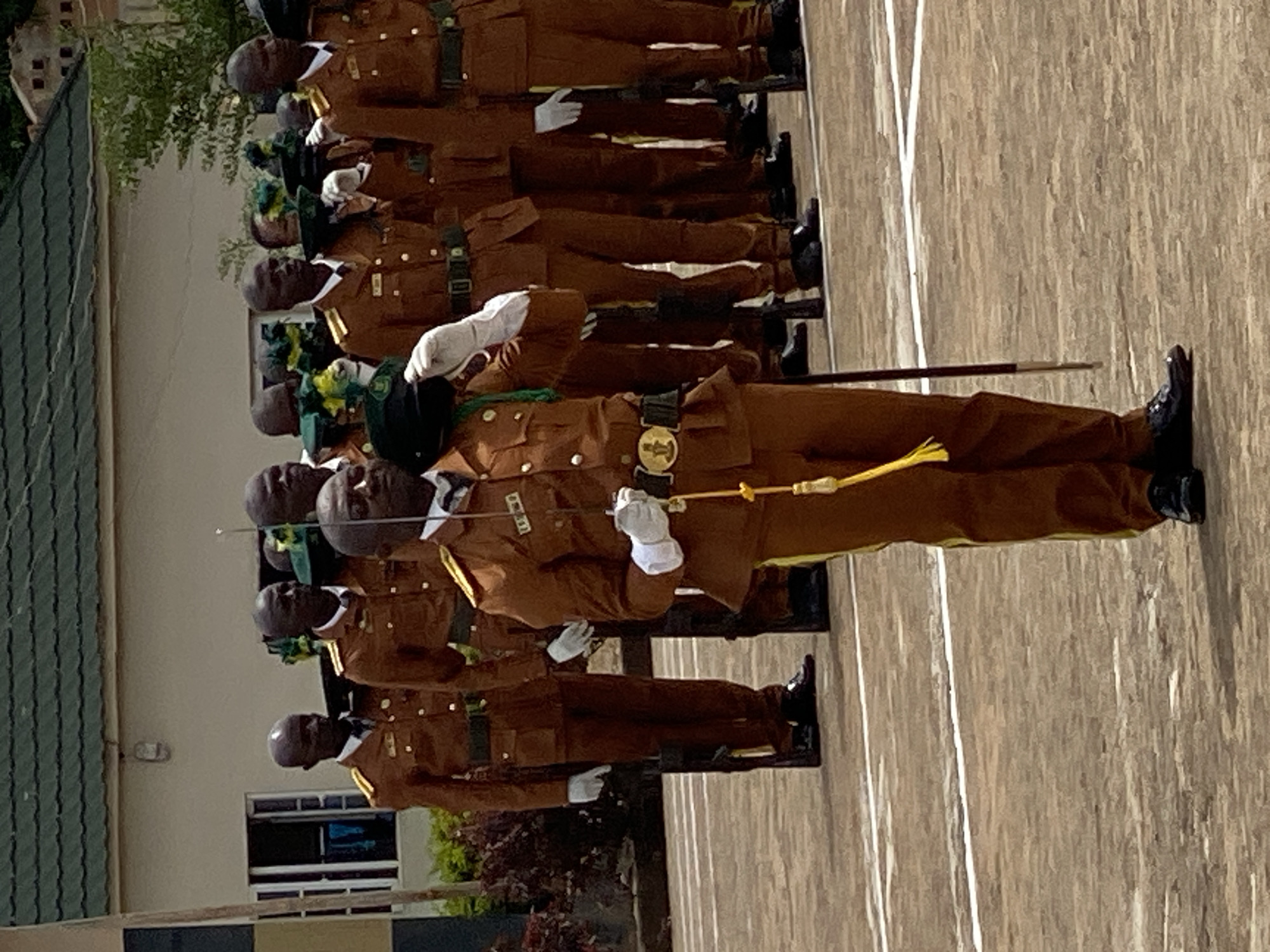 A group of individuals in uniform stand in formation. They are all wearing similar brown uniforms with dark collars and belts, and dark caps. Some are holding swords. The ground they are standing on is a paved surface with white lines. In the background, there is a building with a green roof and some trees. The image appears to be taken during daylight. The people seem to be participating in some kind of formal ceremony or parade, possibly a military or cadet event.