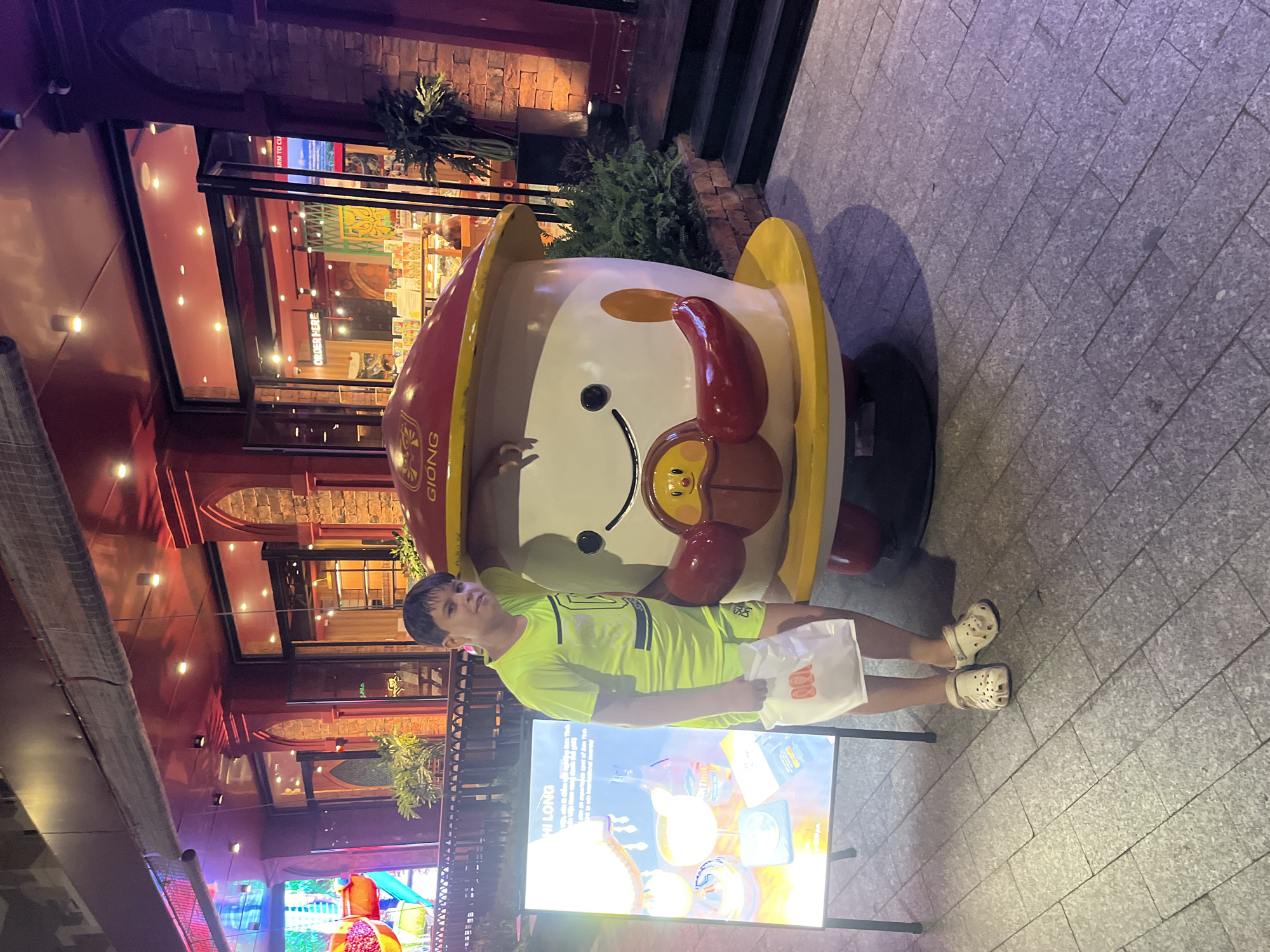 A young boy wearing a neon yellow t-shirt and shorts stands next to a large, cartoonish mushroom-shaped mascot outside a building at night. The mascot has a red cap with "GIONG" written on it in yellow, and a smiling face with two black eyes, an orange cheek, and a red mouth. It is standing on a yellow circular base. The boy is holding a white plastic bag in his right hand and leaning his left arm on the mascot. He is wearing beige clogs. To the boy's left is a lit-up advertisement board displaying images of drinks and text in Vietnamese. The background shows the exterior of a building with large windows and illuminated interiors, suggesting it is a restaurant or shop. Red and brick-colored architectural details are visible. The ground is paved with gray square tiles. The scene appears to be located in Nha Trang, Vietnam, likely on a street or pedestrian area.