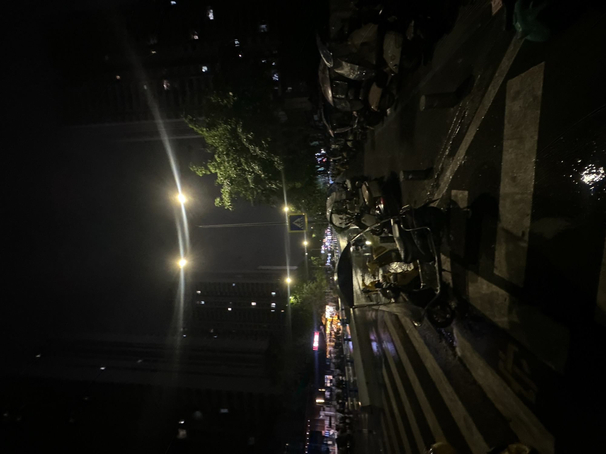 This image displays a nighttime urban street scene in the Chongqing High-tech Zone, China. The ground surfaces are wet and reflective, indicating recent precipitation. Two prominent streetlights in the midground generate intense light and horizontal lens flares. Tall, dark multi-story buildings with scattered illuminated windows are visible in the background on both sides of the street.
In the foreground and extending into the midground, a white-striped pedestrian crosswalk is visible. Along the left side of the street, a row of commercial establishments with illuminated interiors and signage is present. Several motorcycles and scooters are parked in front of these establishments. The right side of the street is occupied by a dense formation of parked covered three-wheeled vehicles, extending into the background. One such vehicle is partially visible in the immediate foreground on the right. Green tree foliage is interspersed along the street, particularly on the right side. A blue pedestrian crossing sign is affixed to a pole in the midground, left of center. No human figures are clearly identifiable or engaged in specific actions within the frame.
