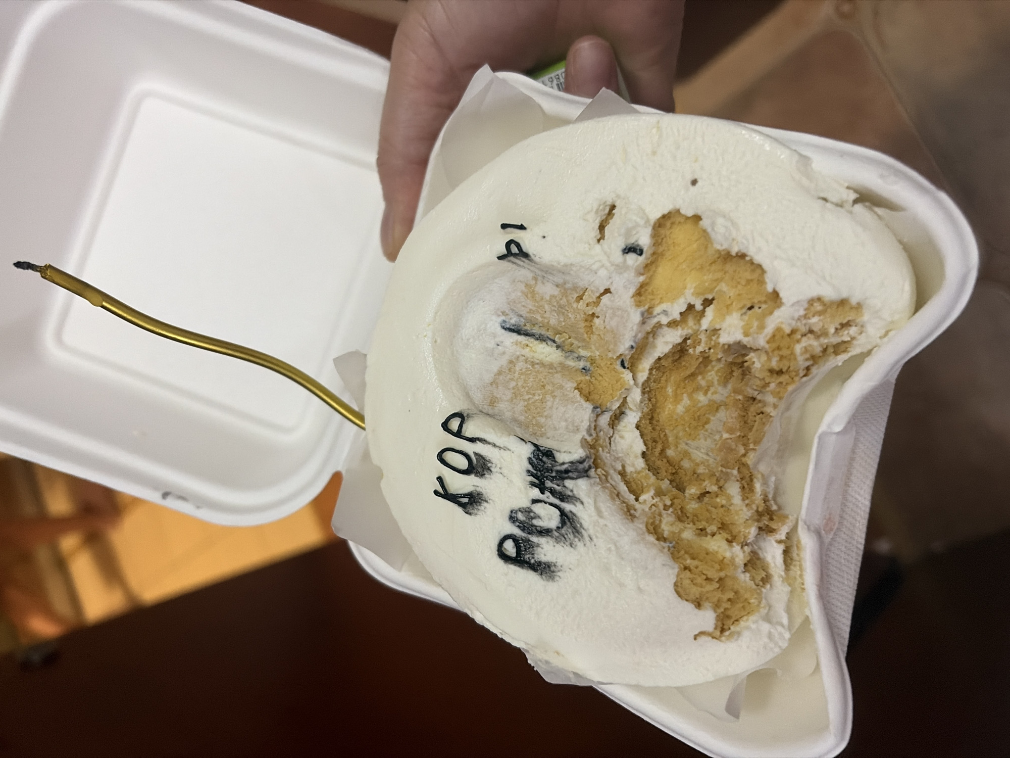 The image shows a partially eaten dessert, possibly an ice cream cake or similar confection, presented in a white takeout container. The dessert is circular and topped with white frosting or cream. Black lettering is visible on the dessert, reading "KOP POR 14". A golden-colored stick, possibly a candle that has been lit and partially burned, protrudes from the top of the dessert. The dessert appears to have been bitten into, with chunks of a lighter colored cake or biscuit base visible beneath the frosting. A hand is holding the takeout container. The background suggests an indoor setting with warm lighting. The location in Budva, Montenegro is not indicated by any visual elements in the image.