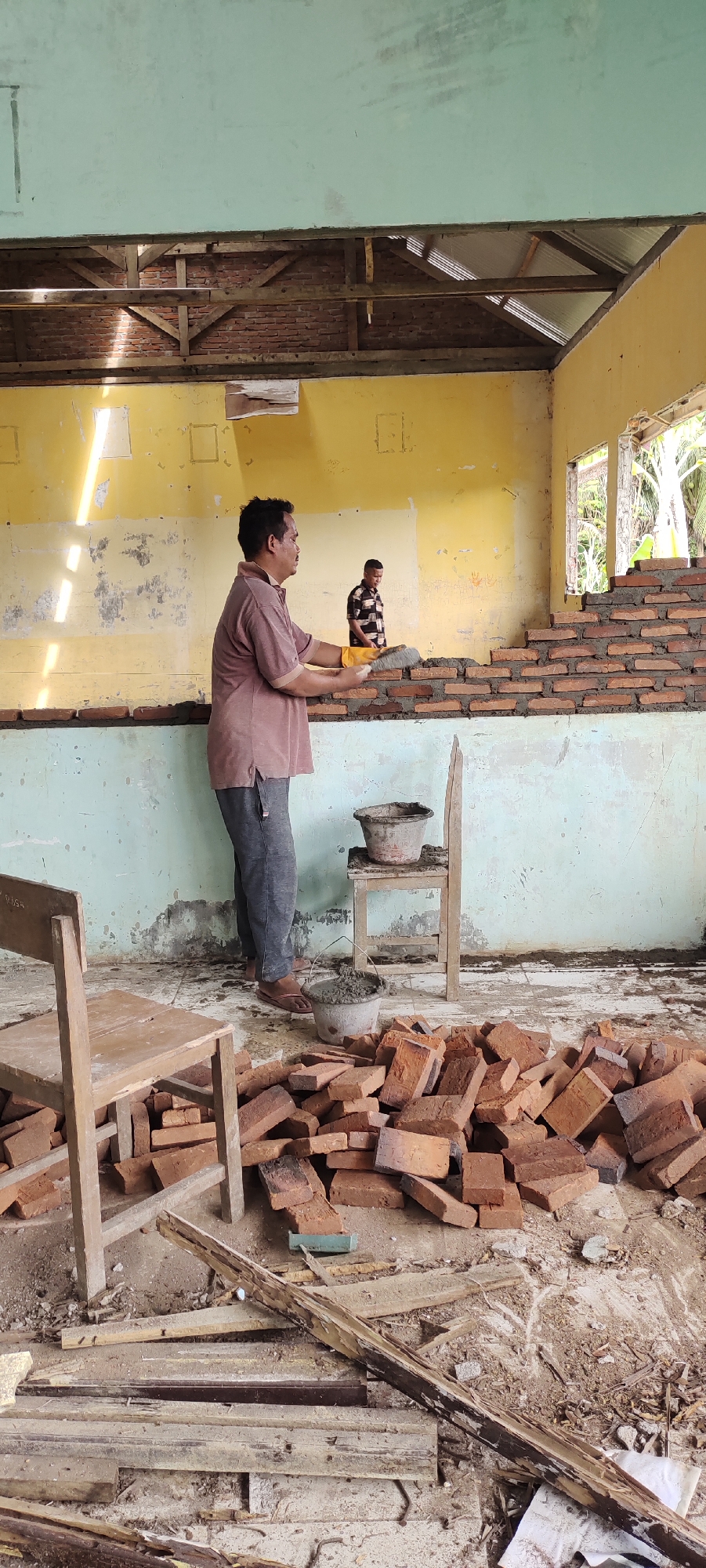 A medium-shot captures a scene in Langnibong, Indonesia, depicting two men engaged in bricklaying inside what appears to be a dilapidated building. One man, facing left, is actively working on the brick wall, wearing a reddish-brown shirt, dark pants, and yellow gloves. He holds a trowel laden with mortar and bricks. A second man stands in the background, to the right of the first, wearing a black and white checkered shirt, observing the work.
In the foreground, a pile of bricks rests on the floor, along with debris and scraps of wood. Two wooden chairs are also present, one with a bucket of mortar on it and one next to the brick pile. The walls of the building are partially yellow and partially light blue, with clear signs of age and disrepair. The structure's roof and upper walls are incomplete, revealing the brickwork beneath and allowing natural light to enter. An open window on the right side of the frame reveals foliage outside. The scene suggests an ongoing construction or renovation project within a building in need of repair.
