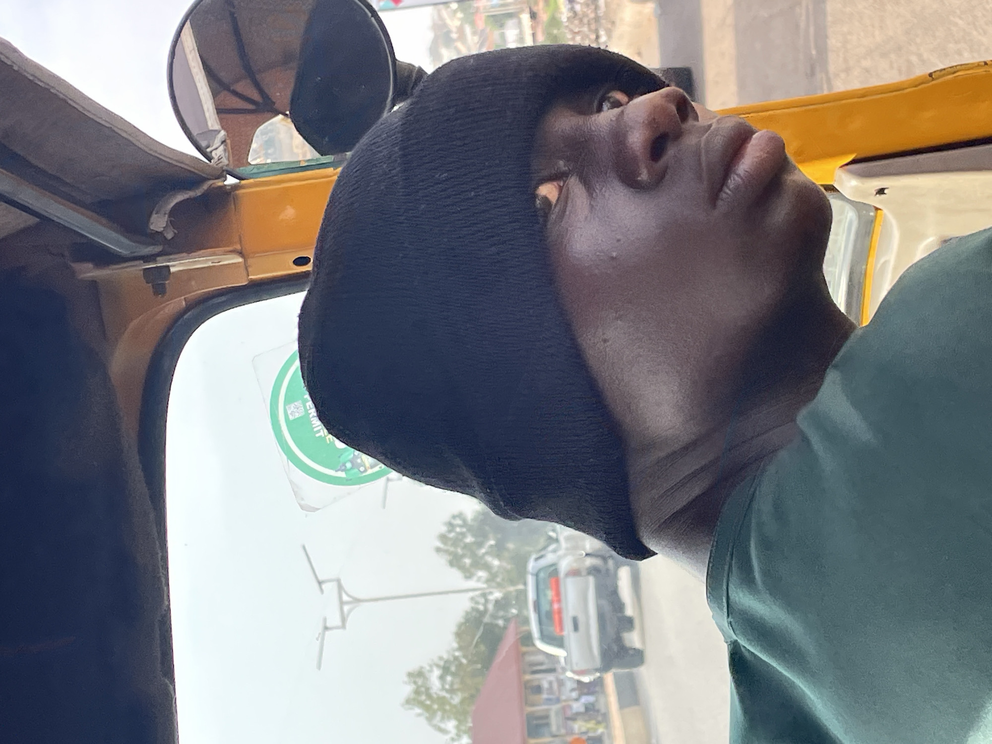 A man in a black beanie looks upward inside a yellow vehicle, possibly a taxi or "keke," in Jos, Nigeria. A round, green permit sticker is affixed to the windshield. Outside the vehicle, other vehicles and buildings are visible on a sunny day.
