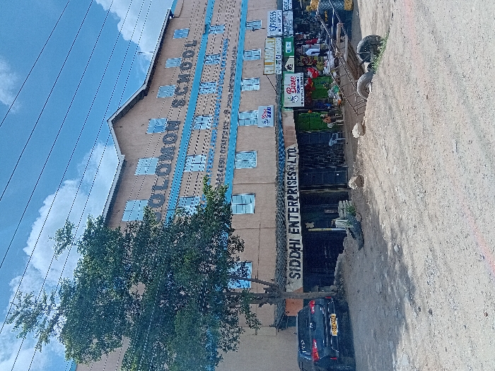 The image shows a street in Mlolongo, Kenya, at the intersection of Kangundo Road and Athi River Road, with a large, two-story building on the left side, with a large sign that reads "Solomon School" on the facade. The building is beige in color with light blue accents and is made of concrete. The building has multiple windows. The building is located at latitude -1.397, longitude 36.942. In front of the building is a large tree and a parking lot. There is a black vehicle parked in the lot. There are a variety of shops in front of the school building. One of these shops, which is made of concrete, has a sign above it that reads "Siddi Enterprises Ltd." There is an empty hand truck in the middle of the street. The street is dirt, with a few cracks and potholes. The sky is blue with a few white clouds. The weather is sunny. There are a few people in the street, mostly in front of the shops. They are all wearing casual clothing. Some are looking at the camera.