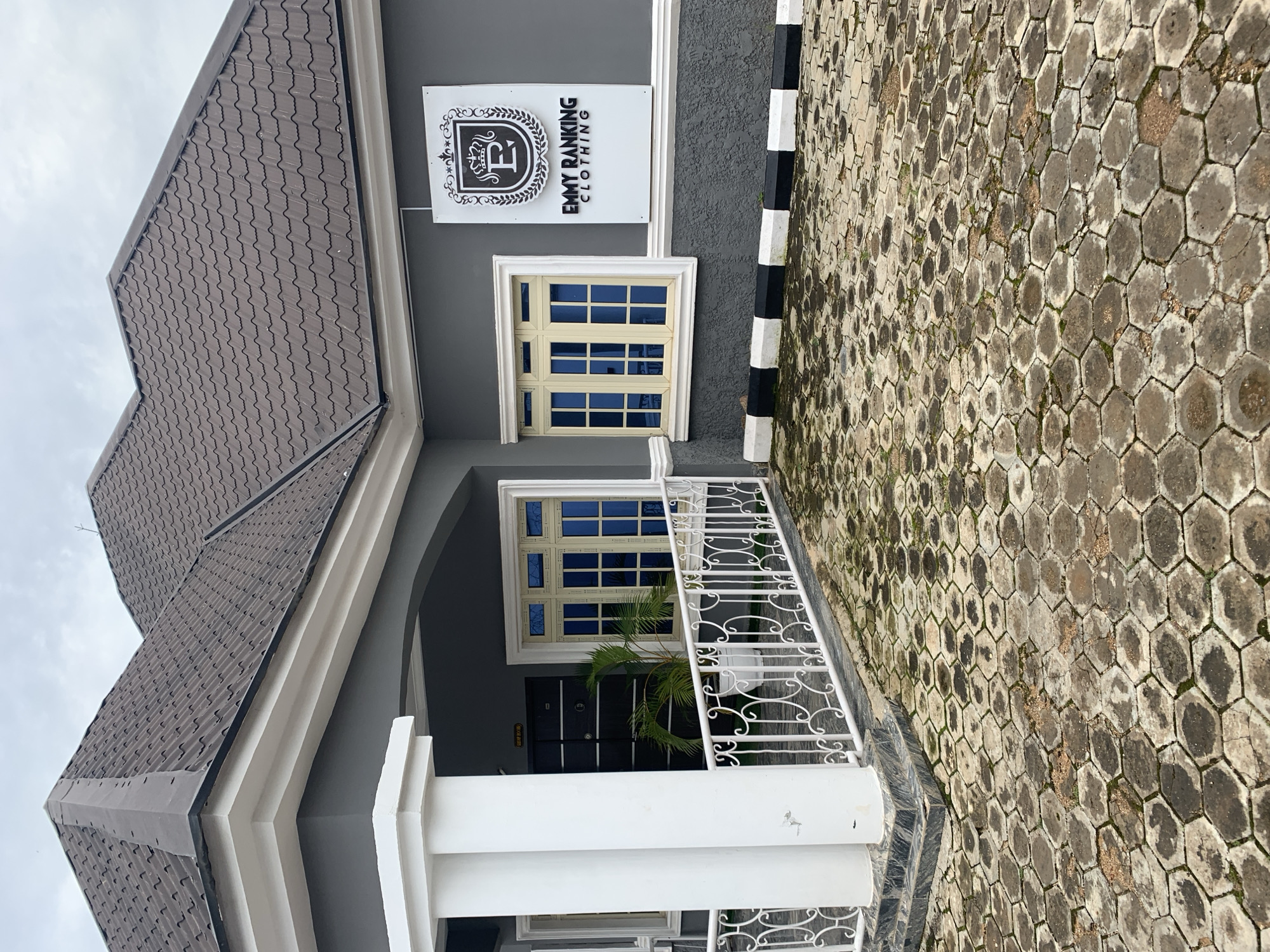 The image shows the exterior of a building with a dark gray facade and a dark brown tiled roof. The building has a prominent white pillar at the front, a white railing, and a paved driveway made of hexagonal stones. On the wall, there is a sign for "EMMY RANKING CLOTHING" with a crest featuring the letter "E". Two windows with light yellow frames and blue glass are visible. A small palm plant in a white pot is located between the pillar and the door. The ground is wet, indicating recent rain. The environment appears to be a street in Gura Top, Nigeria. There are no people in the image.