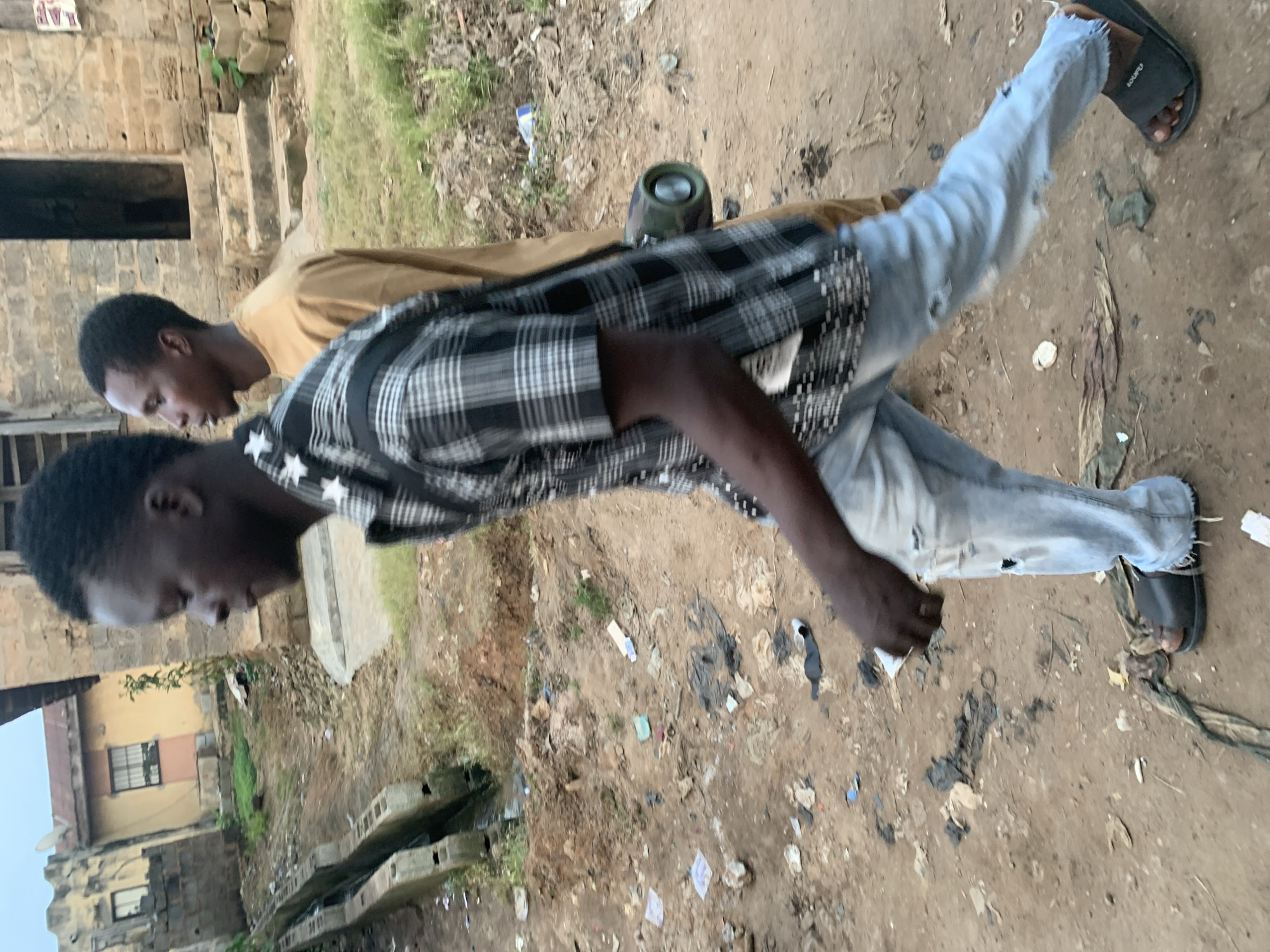 The image shows two young Black men walking on a dirt road littered with debris in Shagamu, Nigeria. The man in the foreground, on the left, is wearing a black and white plaid shirt with white star accents and ripped light-wash jeans, and black open-toed sandals. He is carrying a cylindrical object with a camouflage pattern and metal accents in his right hand, which is slightly extended forward. He appears to be walking towards the right side of the frame, looking ahead and slightly down. The man in the background, on the right, is wearing a tan or light brown long-sleeved garment and is walking behind and to the right of the first man. His face is partially visible, and he is looking forward.
The environment is unpaved and appears to be a side street or path. There are buildings in the background, one with a light-colored facade and a tiled roof, and another made of concrete blocks. A drainage ditch with concrete edges runs along the left side of the path, and its surface is covered with water and debris. The ground is a mixture of dirt, sand, and various types of refuse, including plastic bags, paper, and other unidentifiable items. The overall scene suggests a somewhat neglected urban or peri-urban setting. The lighting indicates it is likely late afternoon or early morning, with diffused light.