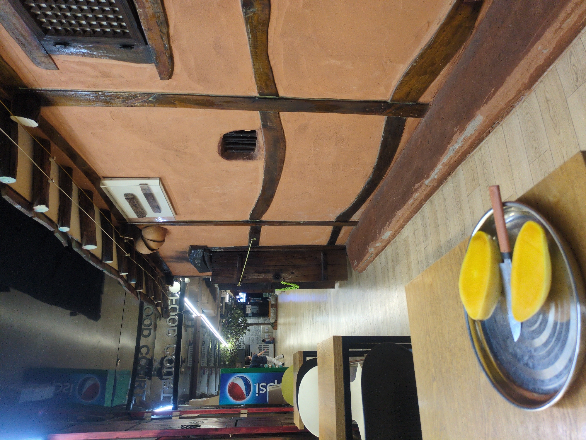 The image depicts the interior of a building in Seoul, South Korea. The ceiling is a traditional Korean style with wooden beams and terracotta-colored plaster. A metal plate with sliced mangoes and a knife sits on a wooden table. Through an open doorway, a street scene is visible with a Pepsi-branded cooler, a Food Court sign, and pedestrians.