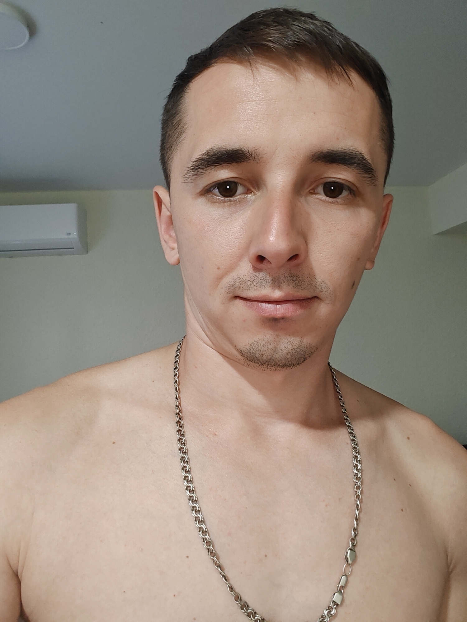A close-up, chest-up, selfie-style photograph shows a light-skinned man with short, dark hair and a small amount of facial hair. He is looking directly at the camera with a neutral expression. He is wearing a silver chain necklace. The background is a plain, light-colored wall with a white air conditioning unit mounted high on the left side. The lighting appears to be from above and to the left. The scene is indoors.