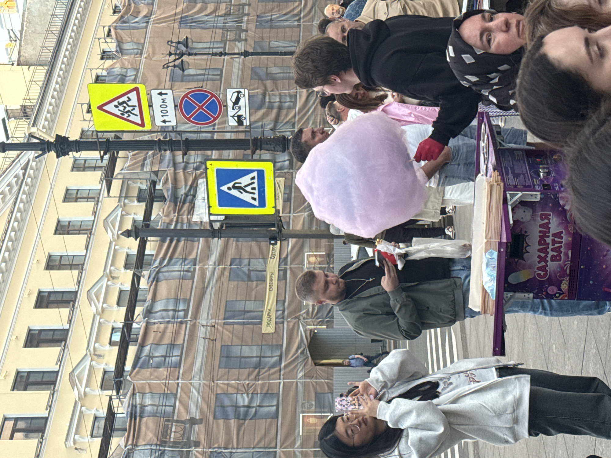 A street scene in Saint Petersburg, Russia, features a woman in a grey hoodie holding up her phone to take a picture of a large pink cotton candy. Beside her, a man in a green jacket looks down at his phone. A street vendor with a red glove is holding the cotton candy stick, offering it to a person in a black hoodie. In the background, a building undergoing renovation is covered in brown scaffolding netting. Several street signs are visible, including a pedestrian crossing sign, a children crossing sign, and a no parking sign. The street appears to be paved, with a crosswalk visible. The lighting suggests it is daytime.