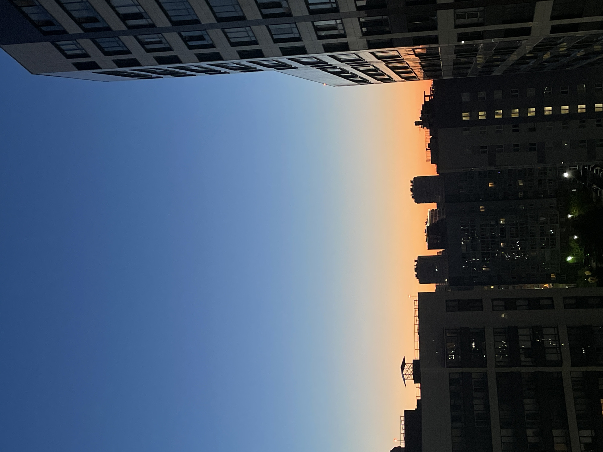 A cityscape at dusk is captured from an elevated viewpoint. A tall, modern apartment building with a dark facade and numerous windows dominates the right side of the frame, angled towards the viewer. In the background, a series of silhouetted apartment buildings of varying heights are visible, their windows beginning to illuminate as the natural light fades. The sky transitions from a deep blue at the top to a soft orange and yellow gradient near the horizon, indicating the setting sun. The scene is set in the city of Sinyavino, Russia, and no people or specific actions are discernible.